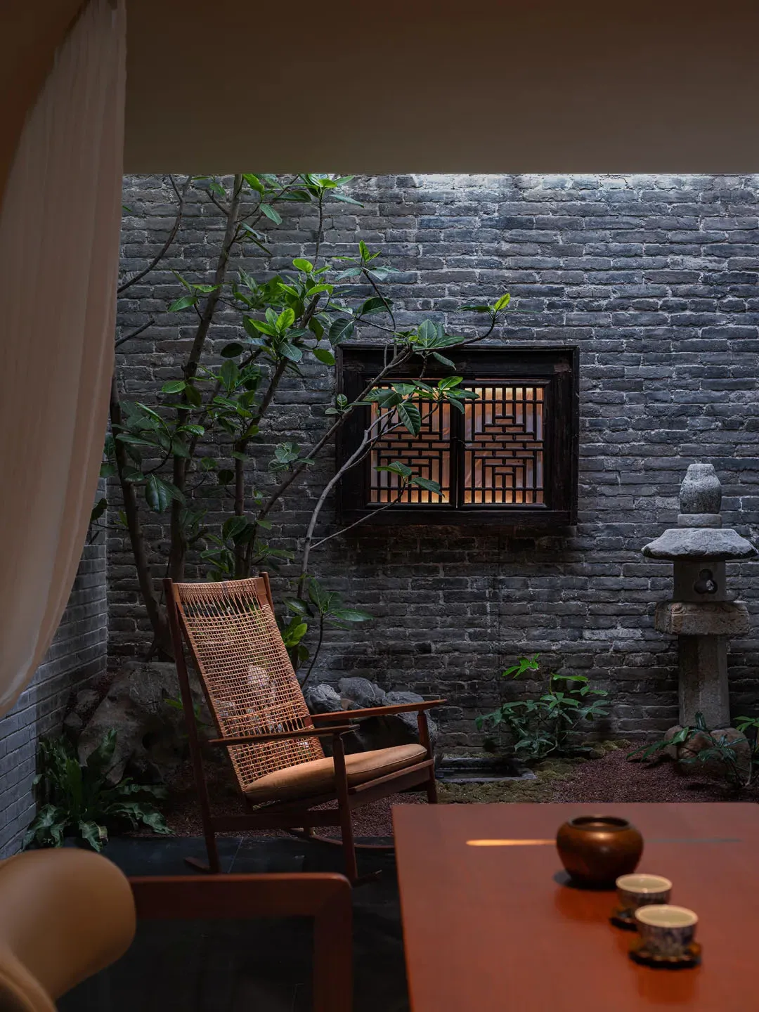 Cozy living room with a brick wall, stone floor, a chair, a dining table, and a potted plant, lit with dim lighting