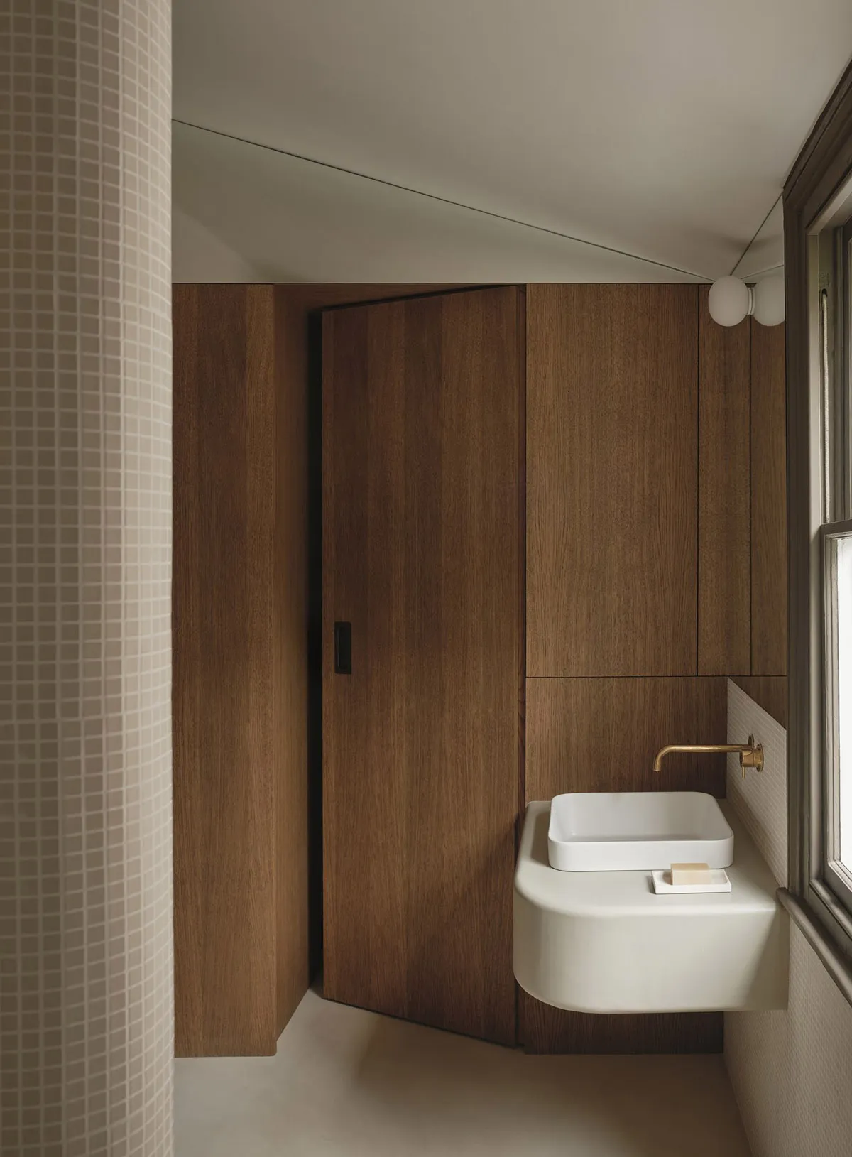 一間現代的浴室，水槽放在窗下，門是棕色的木門，受到自然光的照射
