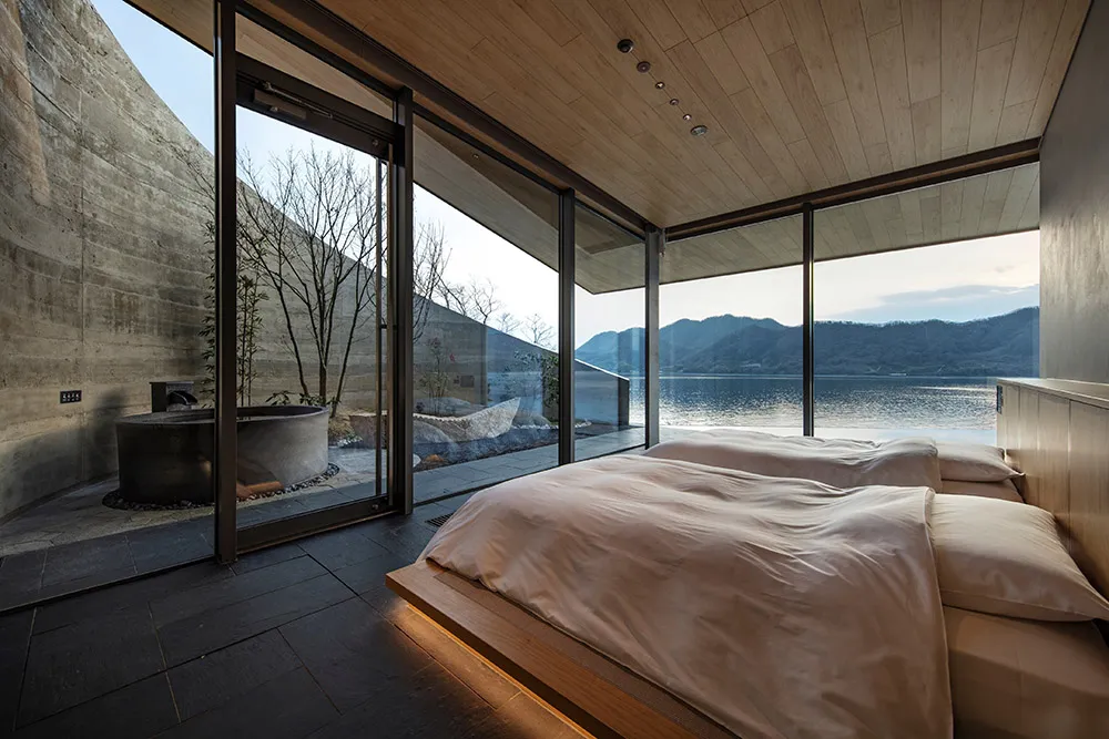 A modern hotel bedroom with a large bed, wooden headboard, and natural light from a large window