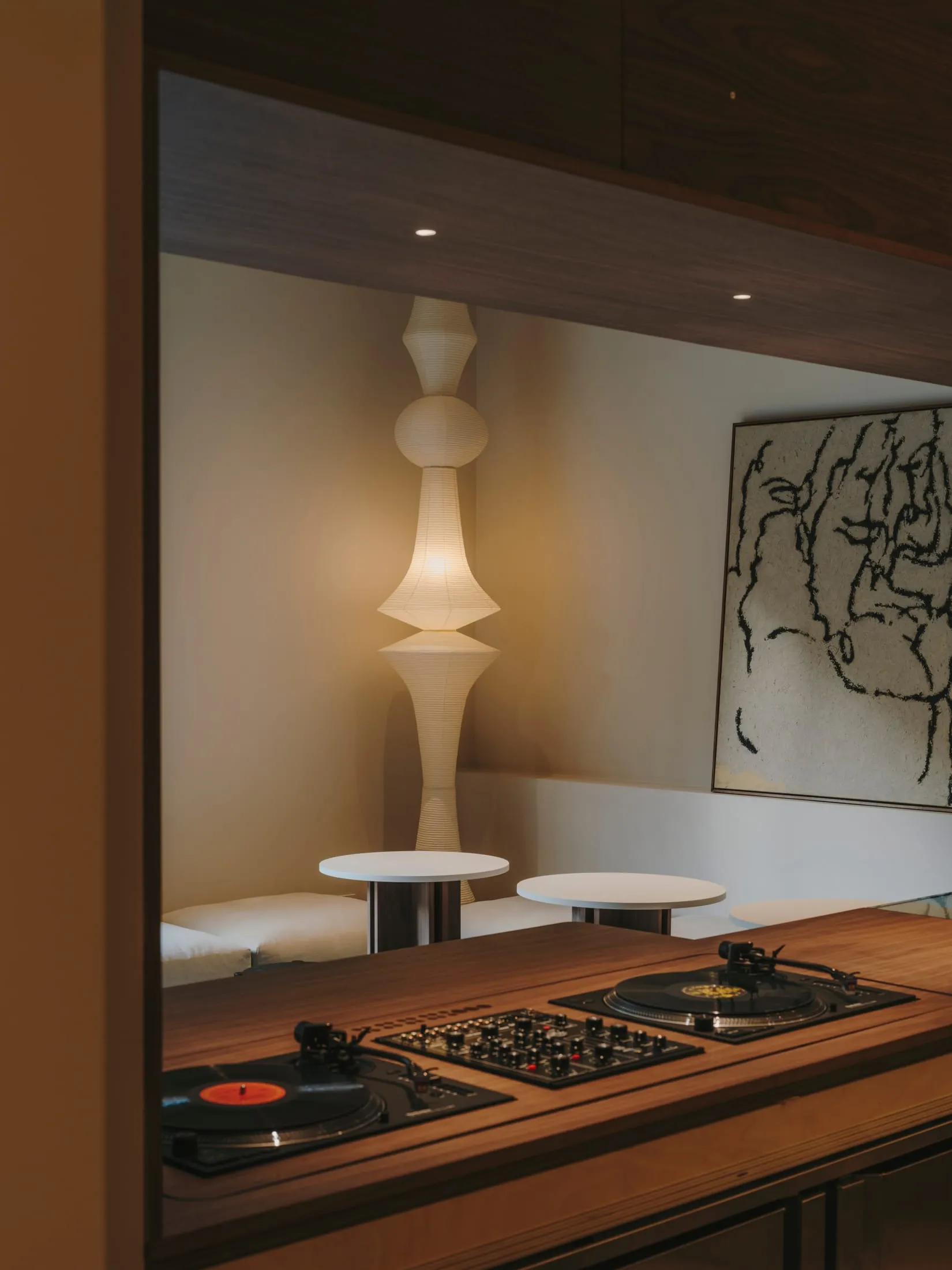 A dimly lit room with a wooden floor, a white wall, and a DJ setup with a turntable and a record player, accompanied by a couch and two chairs