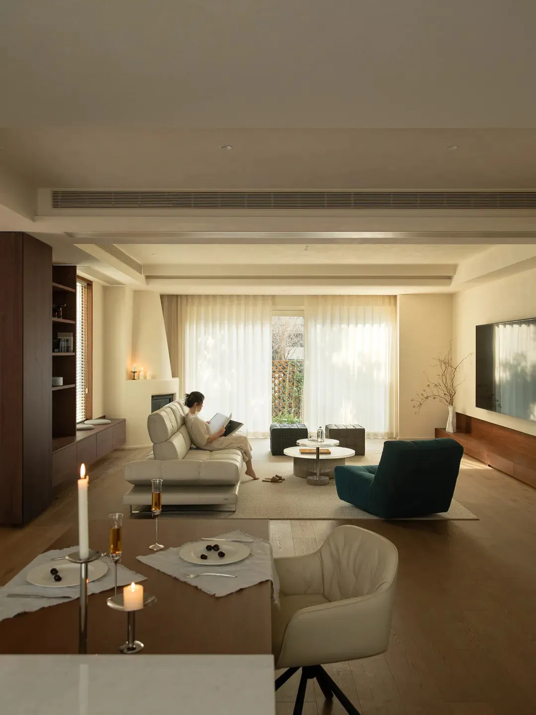 A well-lit living room with a woman on a couch, a dining table, chairs, and a TV, surrounded by white furniture and natural light