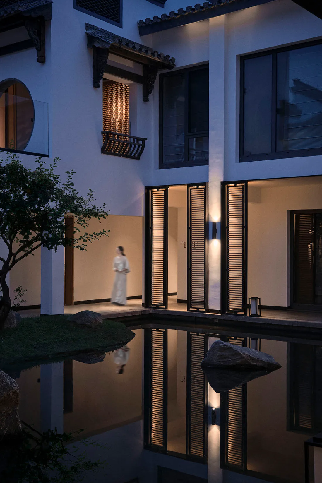 A woman walks in front of a modern building with a reflecting pool in a serene courtyard setting