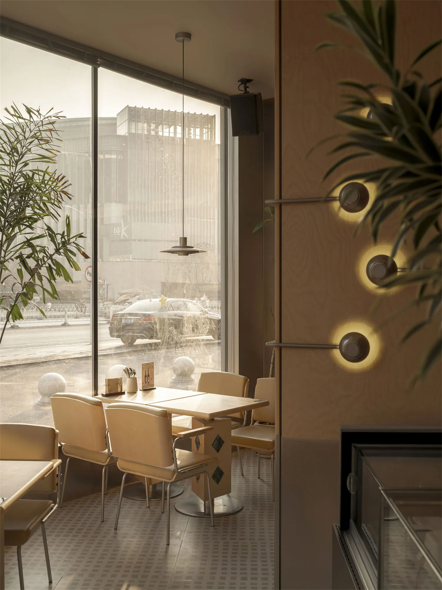 A modern restaurant with a large dining table surrounded by chairs, a potted plant, and a large window, creating a cozy atmosphere with dim lighting
