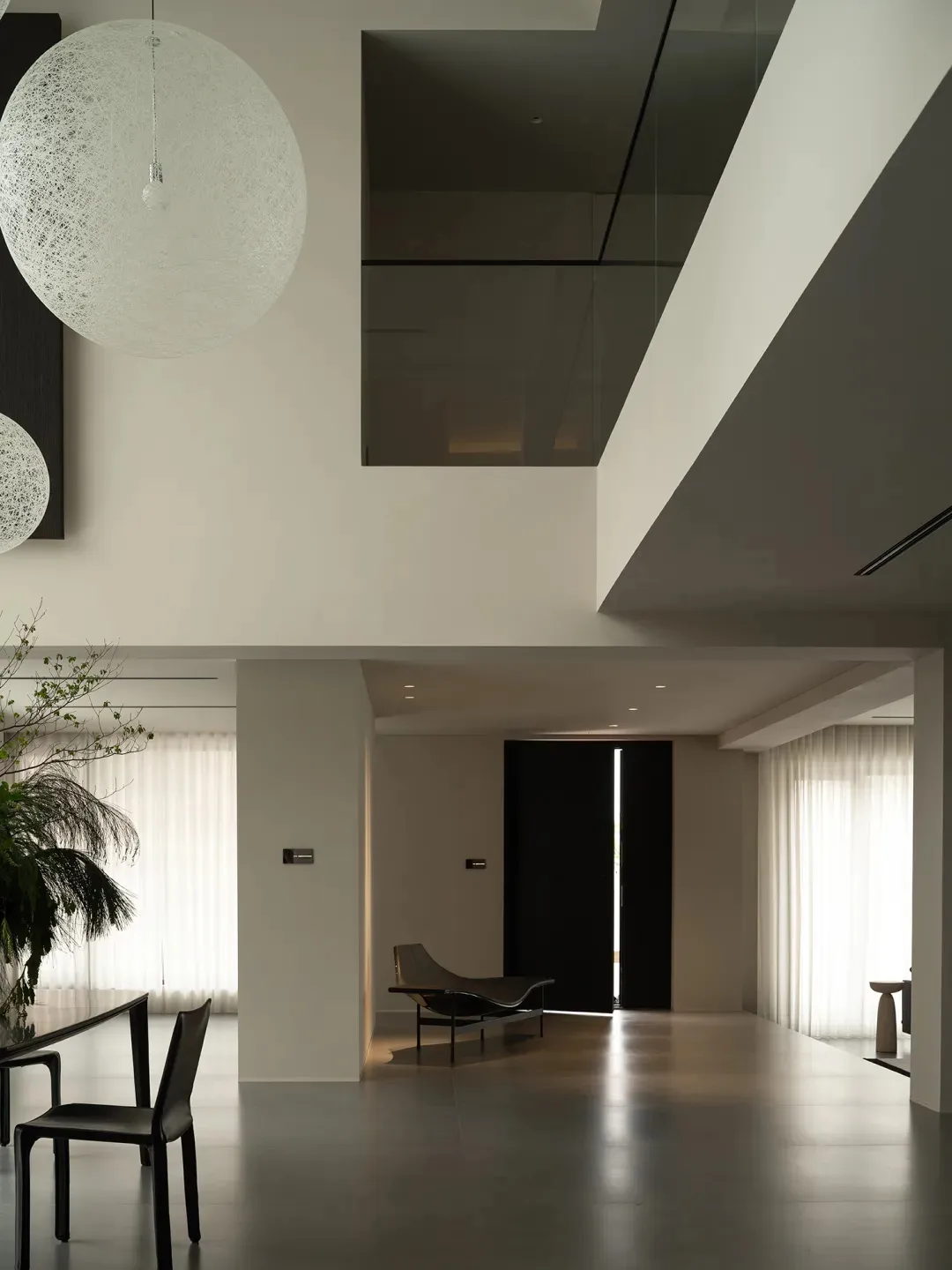 A modern living room with a high ceiling, well-lit space, and various furniture pieces, including a couch, chair, and dining table, with a potted plant and black door adding to the space