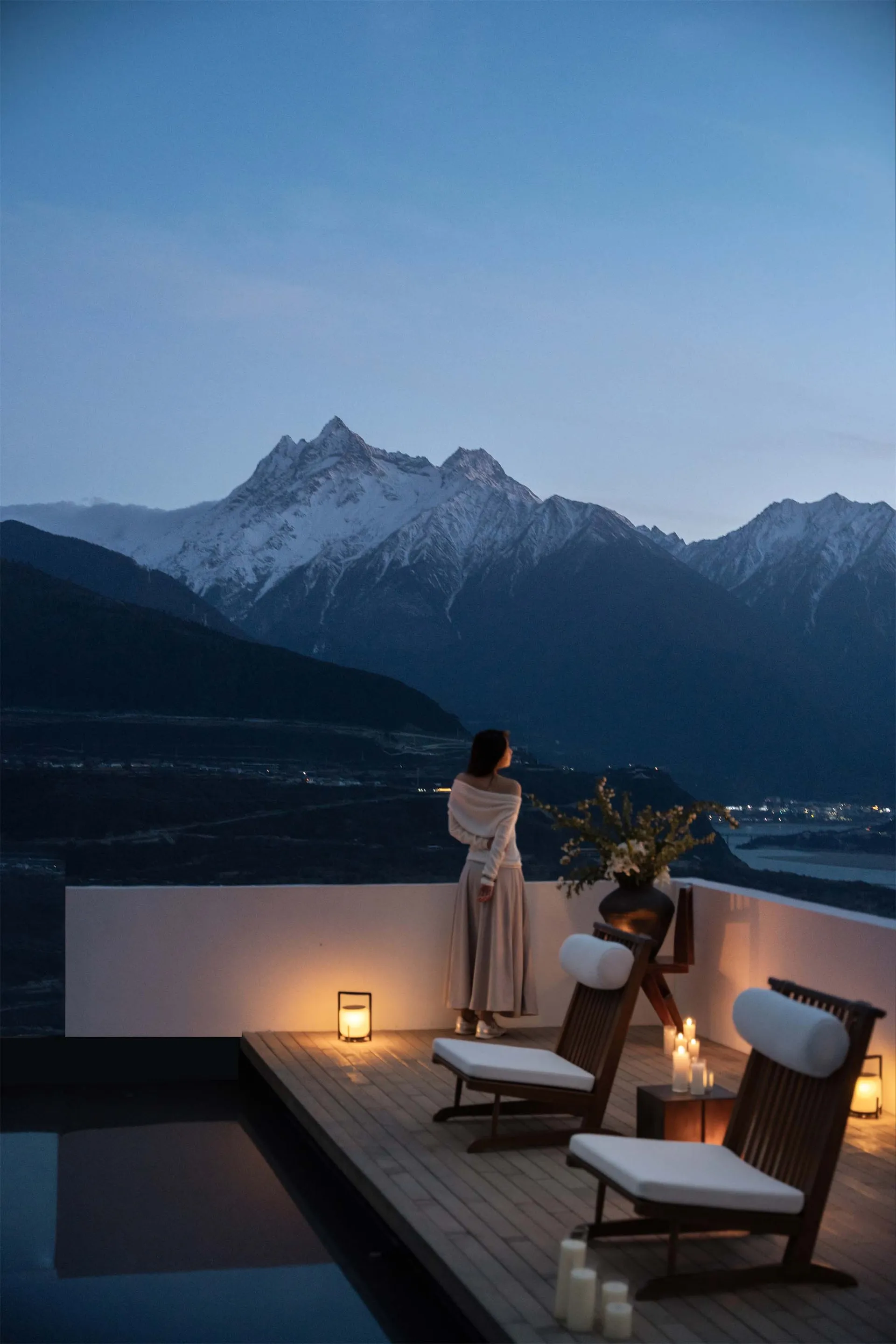 A woman stands on a deck overlooking a mountain range, surrounded by natural scenery and a potted plant, with two chairs and a serene atmosphere