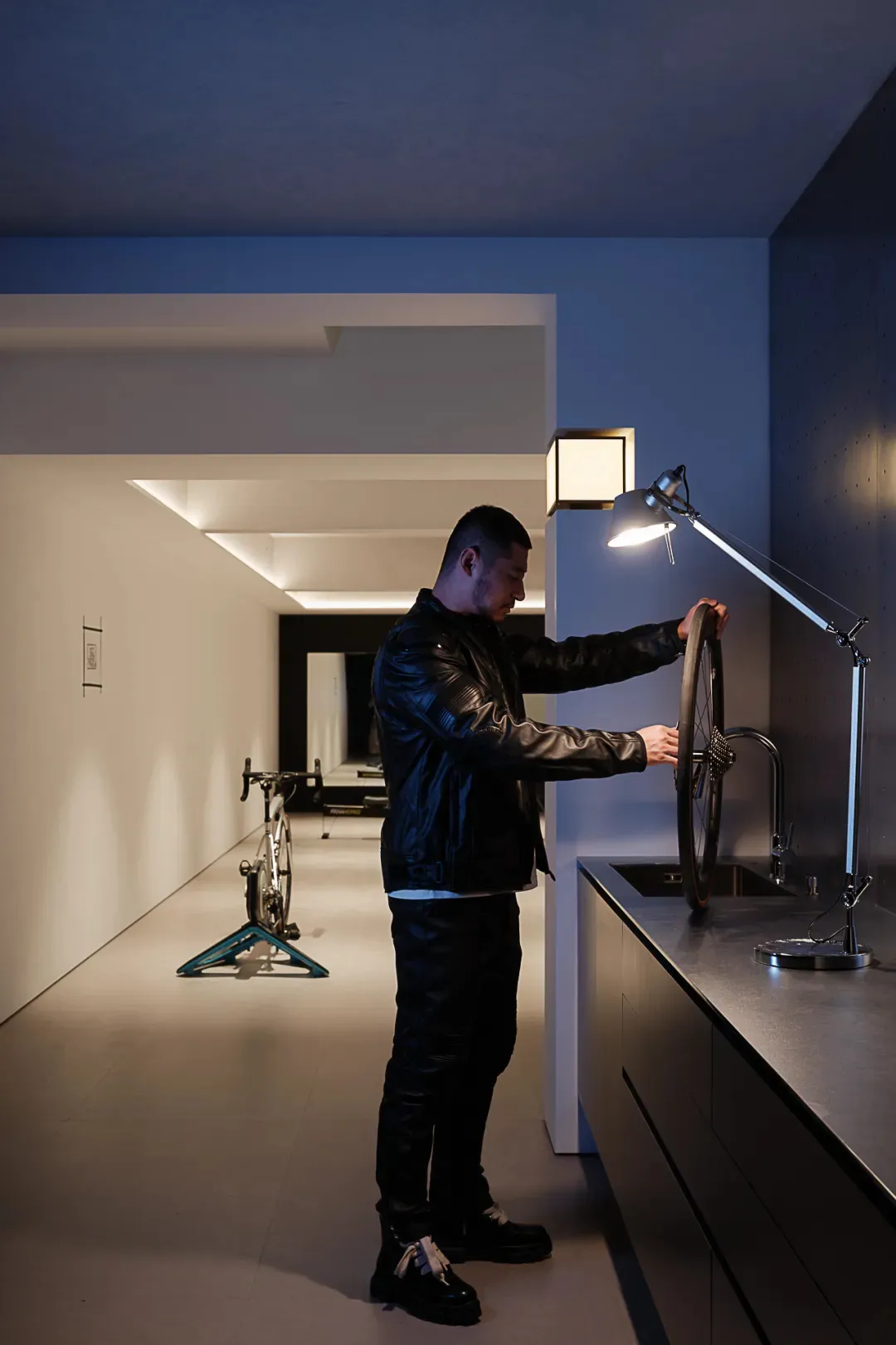 A man in a bathroom, standing next to a sink and a bicycle, with a toilet in the background, wearing a leather jacket and adjusting a bike wheel, in a white-colored room with a light on the wall