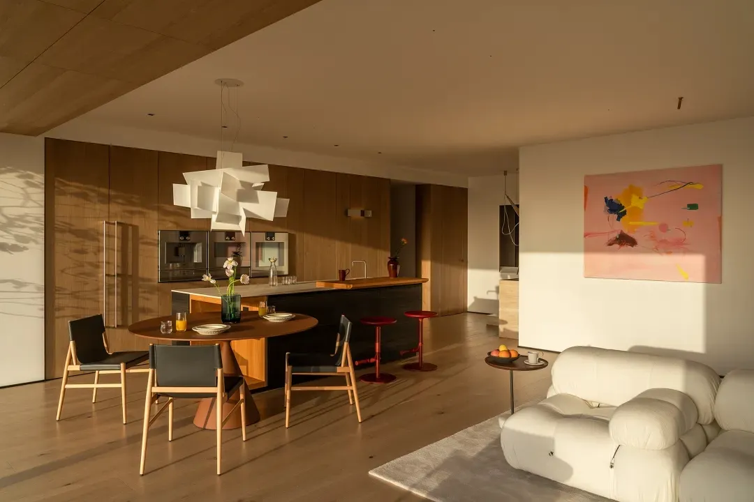 Spacious living room with a wooden floor, a dining table, kitchen area, and natural light from windows, decorated with a vase, potted plant, and bowl