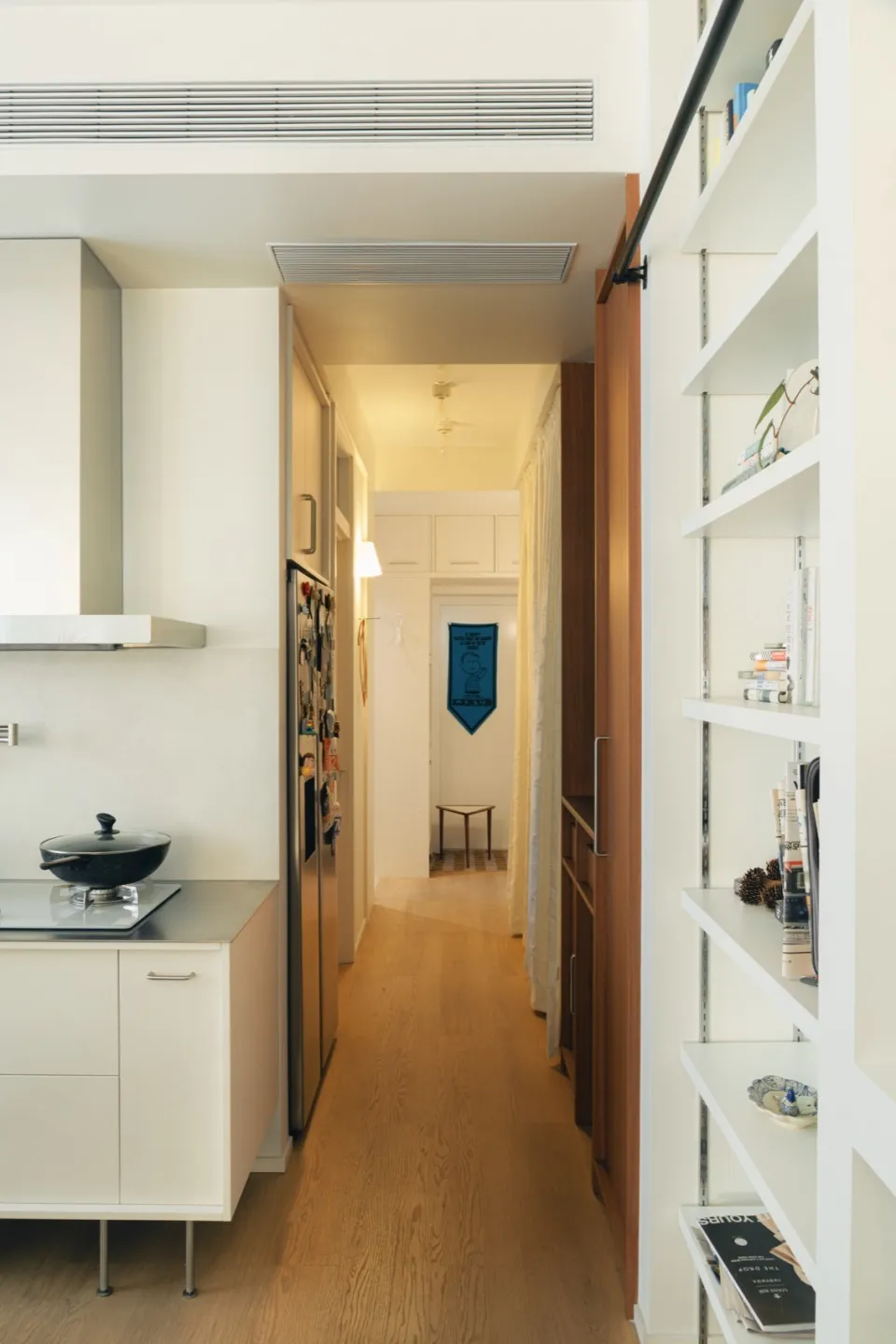 A predominantly white kitchen and hallway with a refrigerator, sink, and bowl on the counter, a doorway, and a flag on the wall, lit by natural light