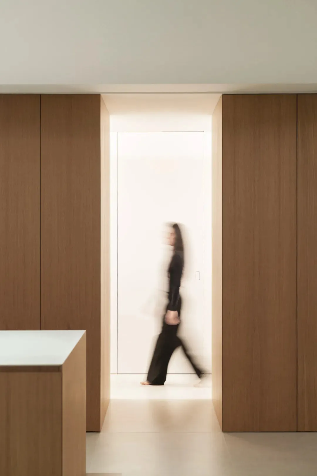 A woman in a black dress walks towards a sink in a kitchen with a wooden floor and a white door