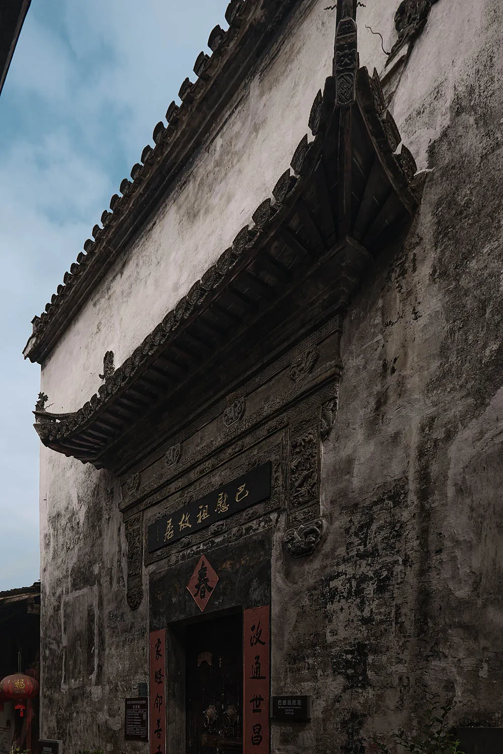 A serene courtyard entrance with a stone building, a red wooden door, and natural sunlight