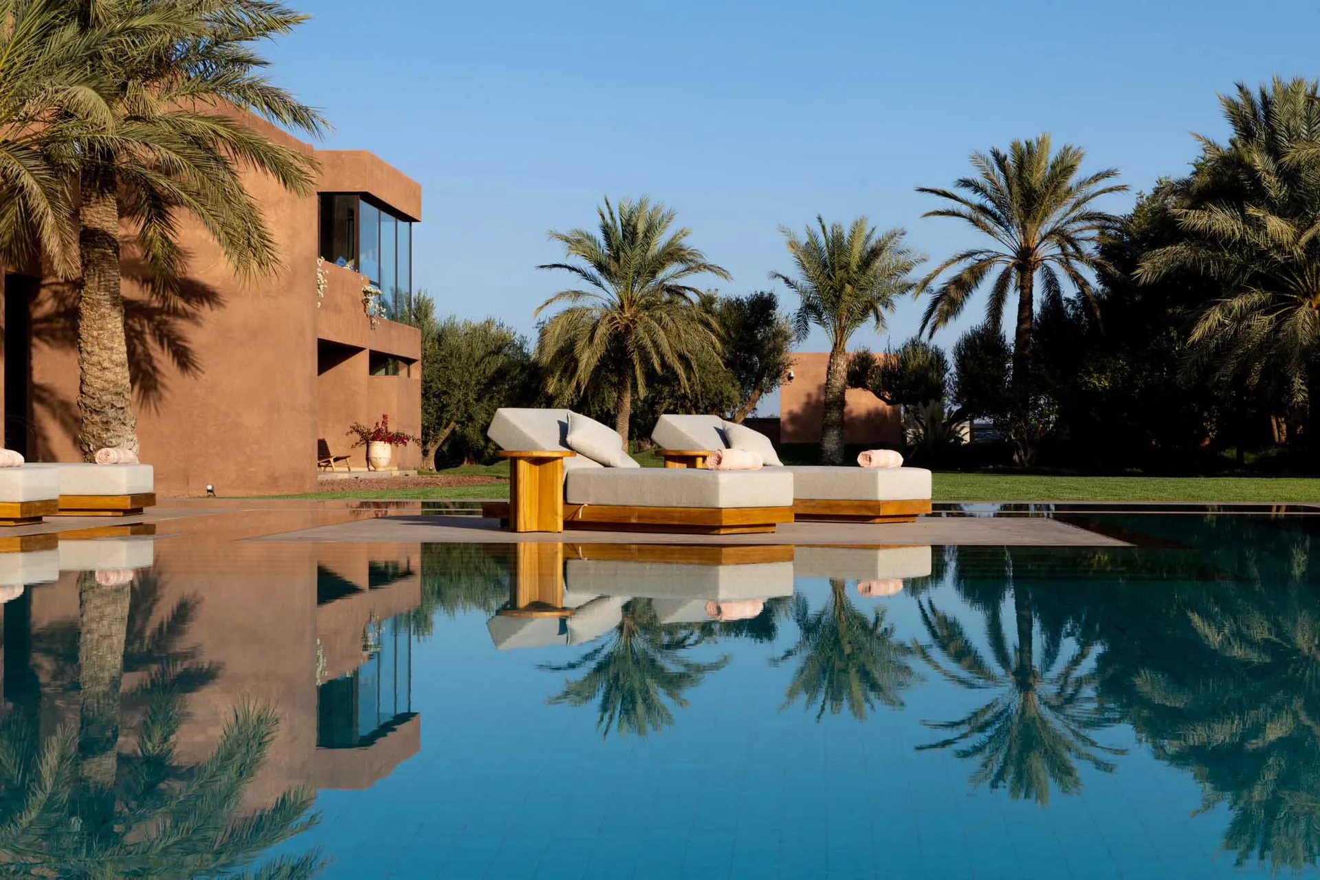 A modern poolside patio with a lounge chair surrounded by palm trees and a clear blue pool