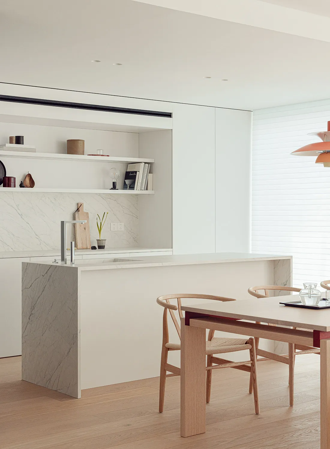 A modern kitchen with a wooden dining table and marble top, surrounded by white cabinets and wooden chairs, with natural light streaming in through the windows