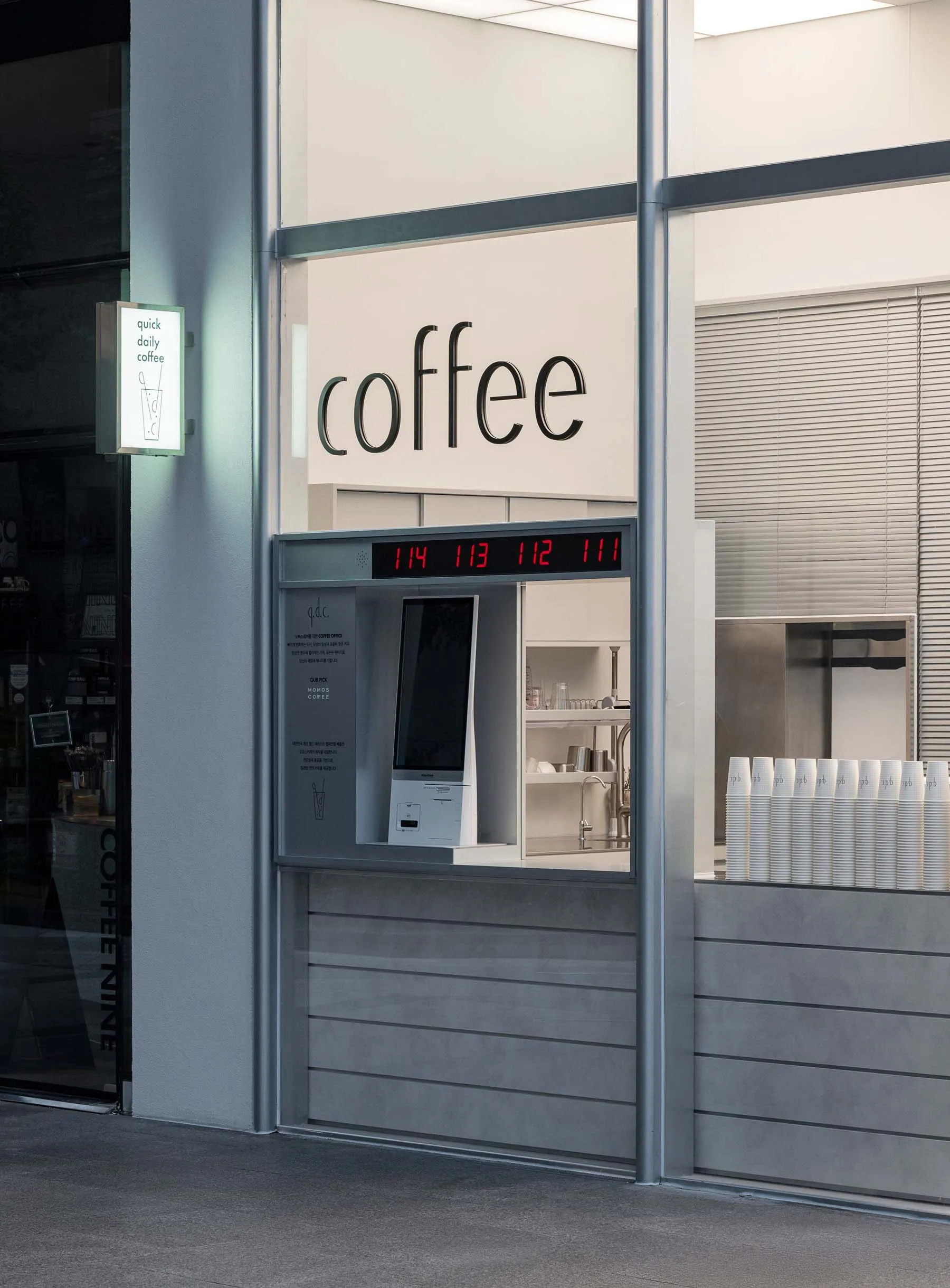 一間現代化的咖啡店，擁有玻璃門面、白牆和光滑的金属柜台，內部擺放咖啡機、杯子和瓶子，配上明亮和熱情的照明
