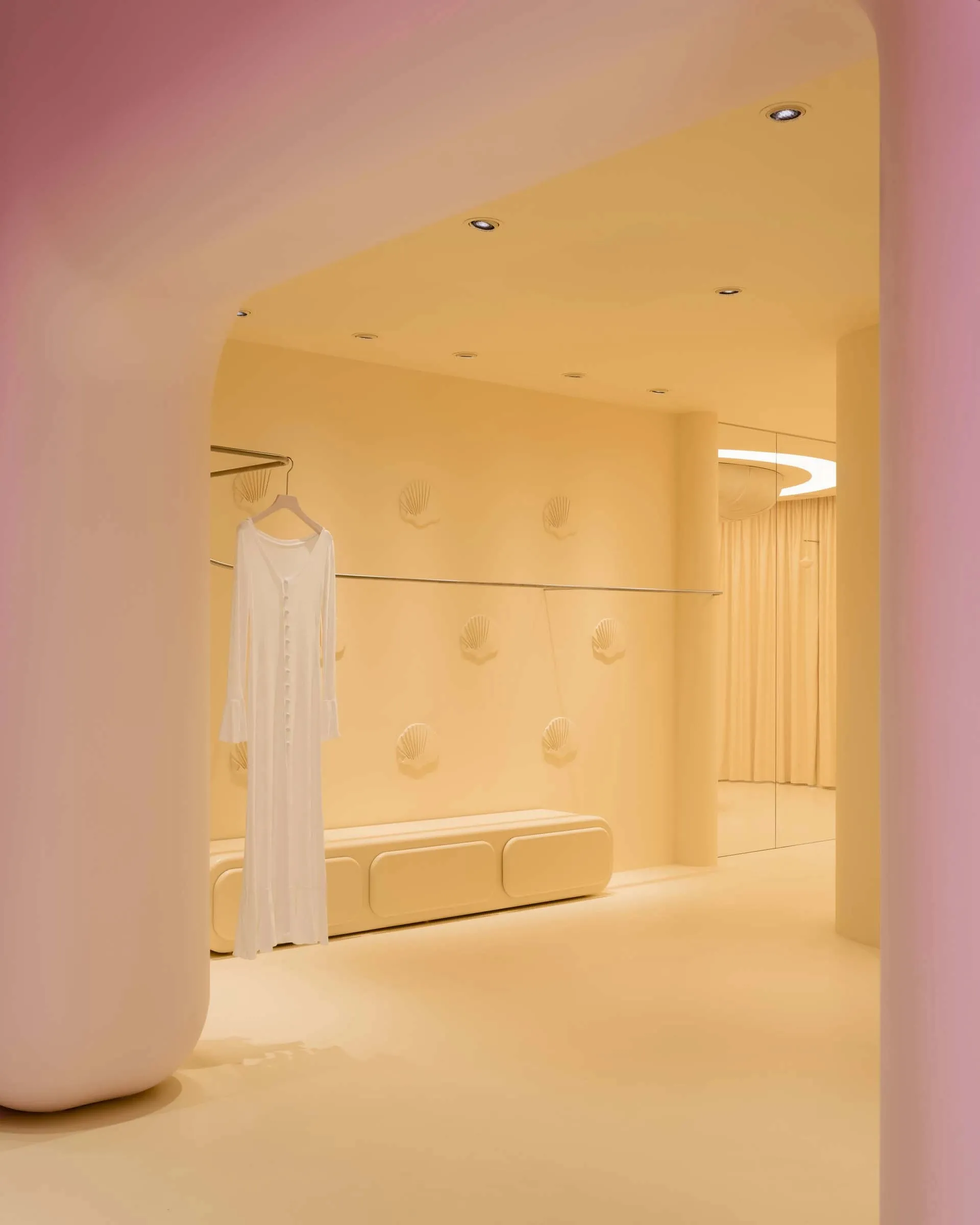 A cozy dressing room with a pink accent wall, a white dress on a rack, and a bench in the center of the space