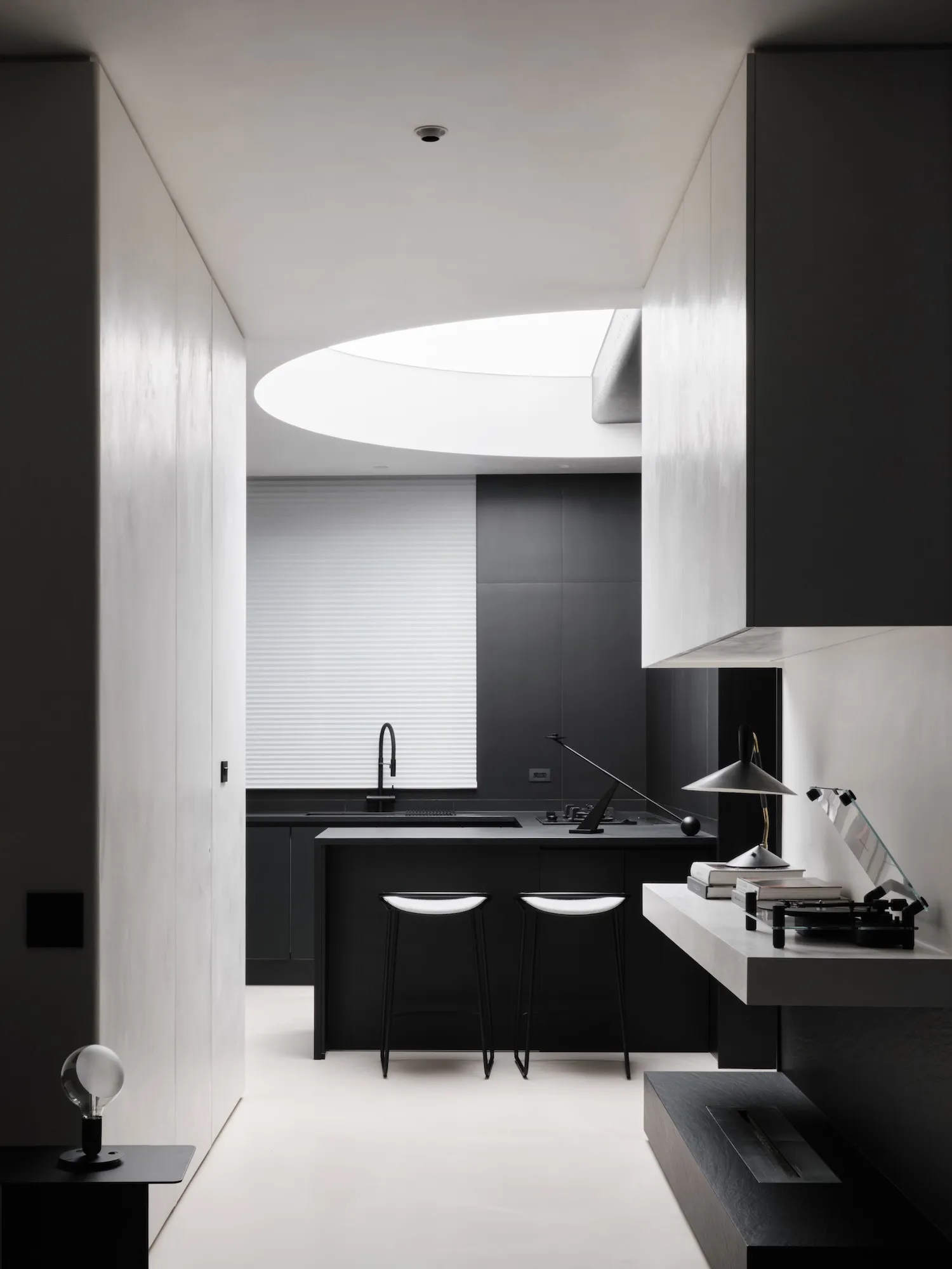 A dimly lit modern kitchen with a black counter and sink under a window, surrounded by three black and white chairs