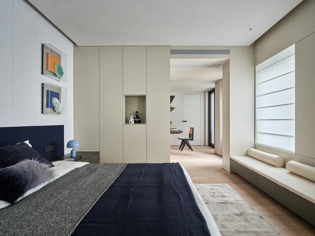 A modern bedroom with a gray color scheme, a blue blanket, and natural light from a window, featuring a bed and a bench in a well-organized and comfortable space