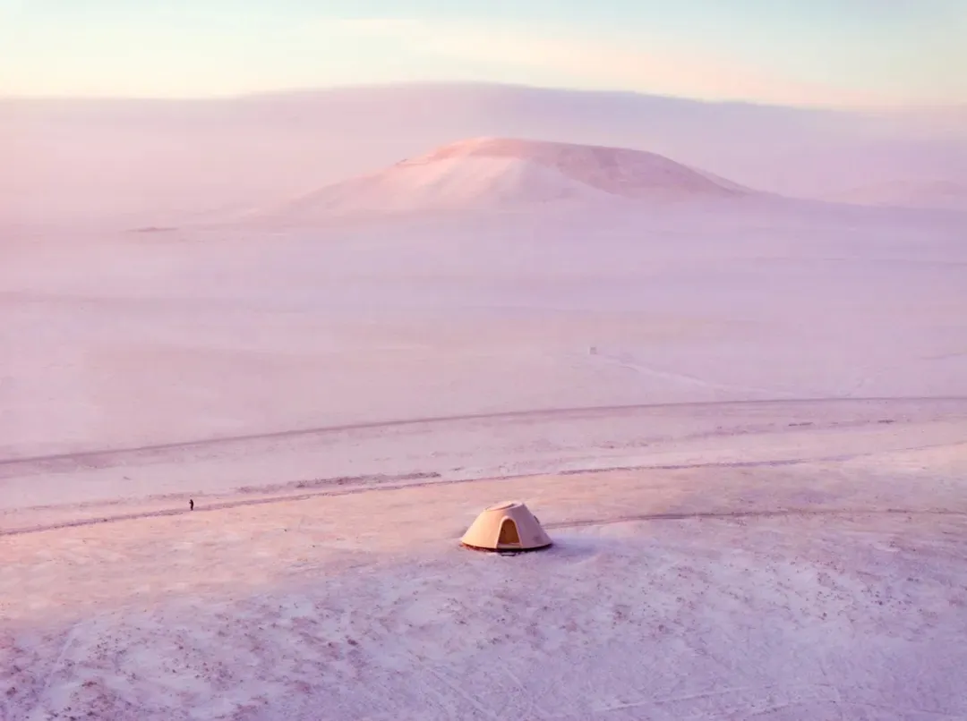 A snowy landscape with a tent in the middle, surrounded by a field and a person standing nearby