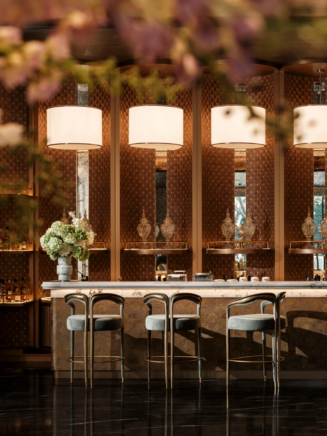 A modern bar with a marble counter, stools, and a vase of flowers, decorated with bottles and cups, and two potted plants, under soft and warm lighting