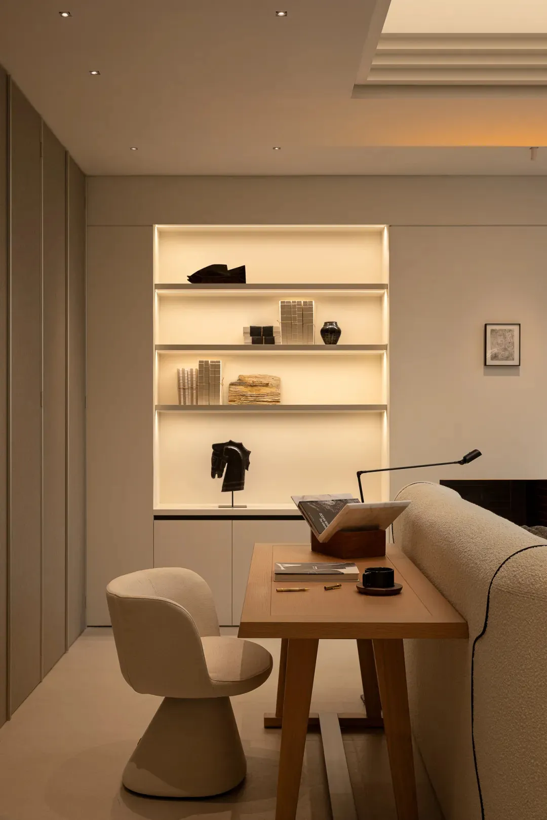 Comfortable living space with a white couch, a wooden chair, and a table, featuring a bookshelf with books and a lamp, well-lit with natural light from a window