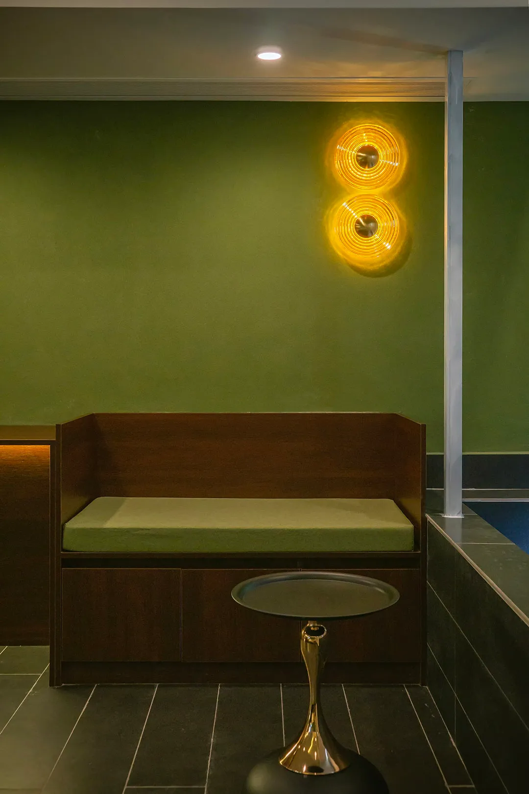 A small, cozy room with green walls, a wooden bench, and a green cushion, lit by a wall light