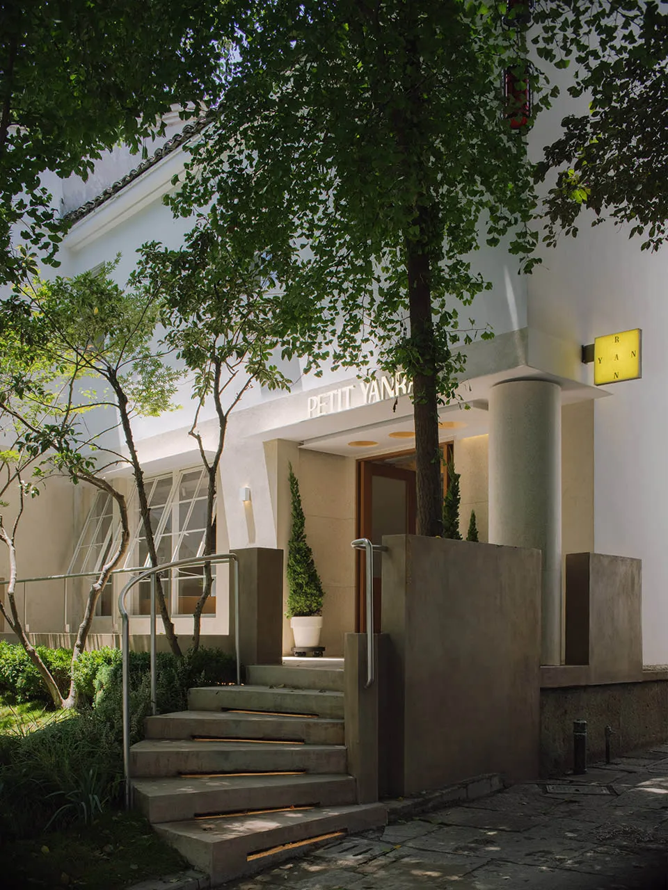 Photograph of a modern building with a white facade, featuring a concrete staircase and a wooden door, under natural light
