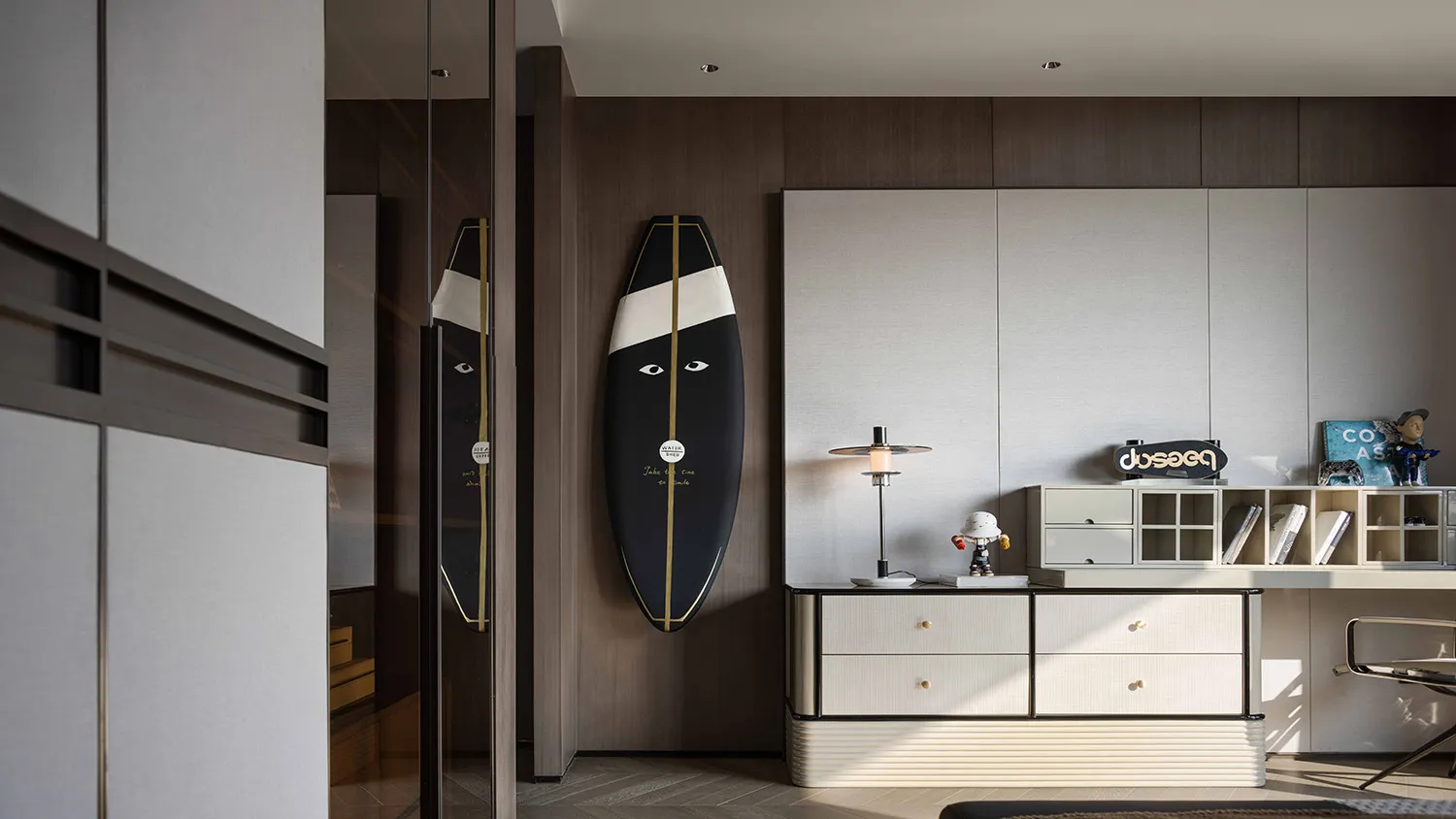 A modern living space with a wooden floor, a desk, a chair, and a black and white surfboard on the wall, with natural light streaming in through the window