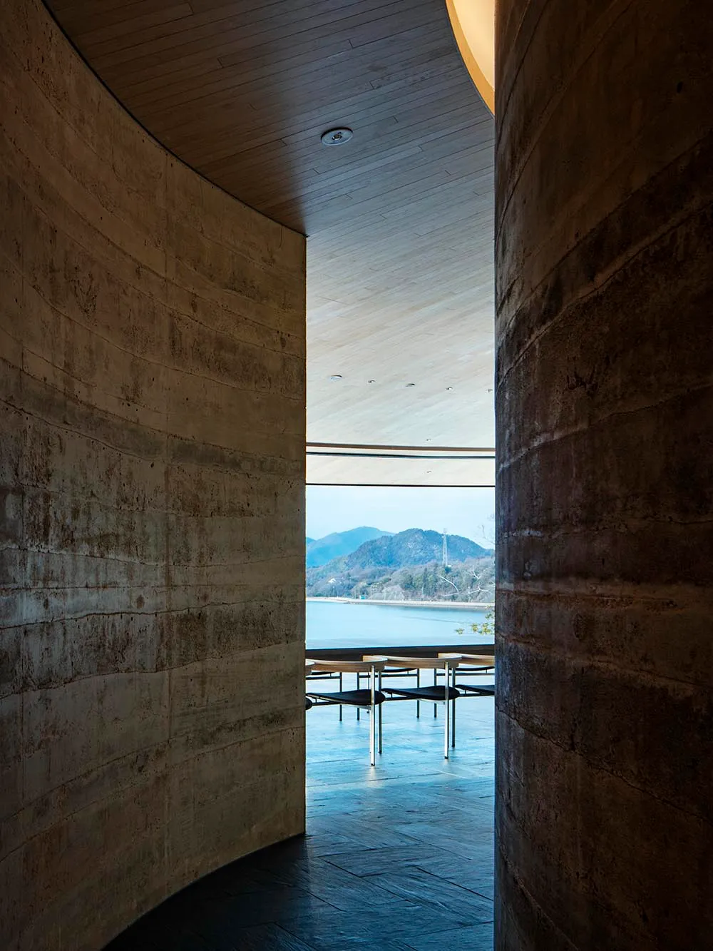 A modern restaurant with a large dining table and chairs, surrounded by a large window offering a view of the water outside, with natural light and a comfortable atmosphere