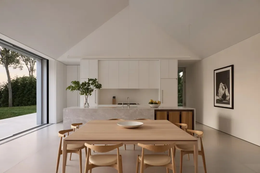 A modern kitchen with a wooden dining table, white cabinets, and natural light from a window, set with a bowl and oranges