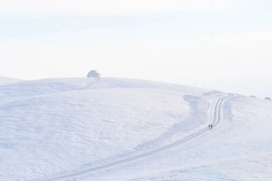 A snowy landscape with a road going up a hill, featuring two people walking and a car in the distance