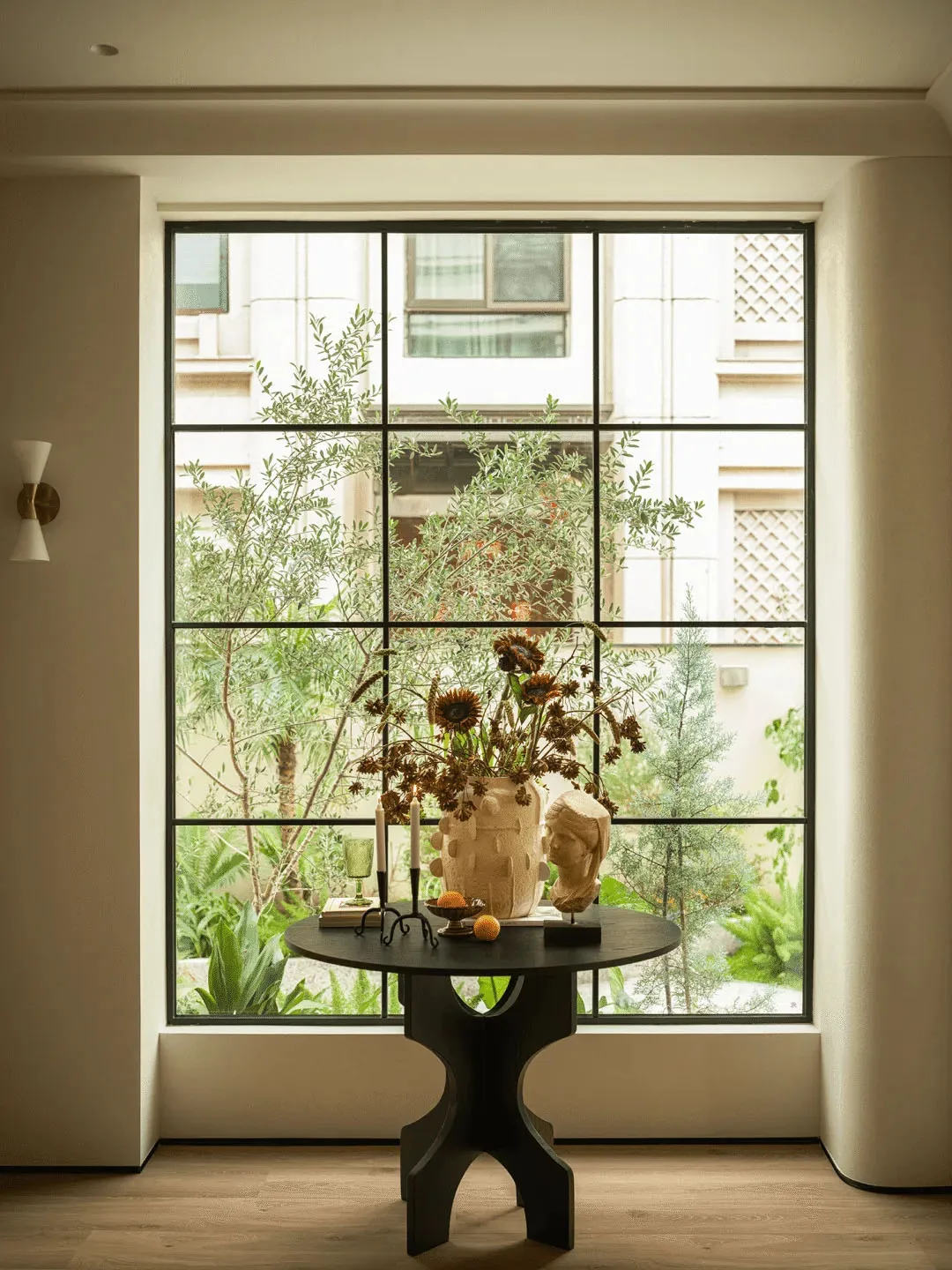 A living room with a table in front of a large window, featuring a vase of flowers and a candle, surrounded by natural light
