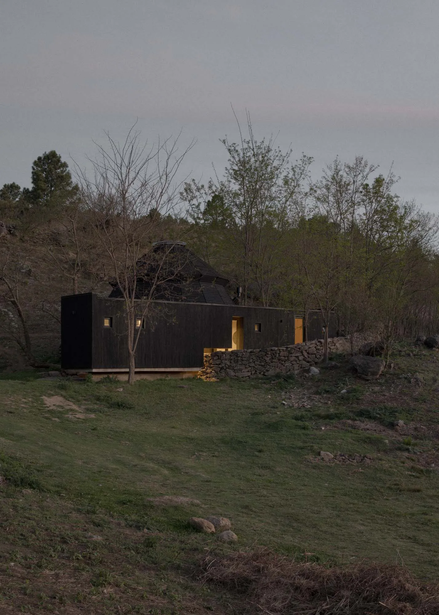A small, dark wooden cabin with a stone wall, surrounded by trees, featuring a mix of a living room and bedroom with a bed and couch inside