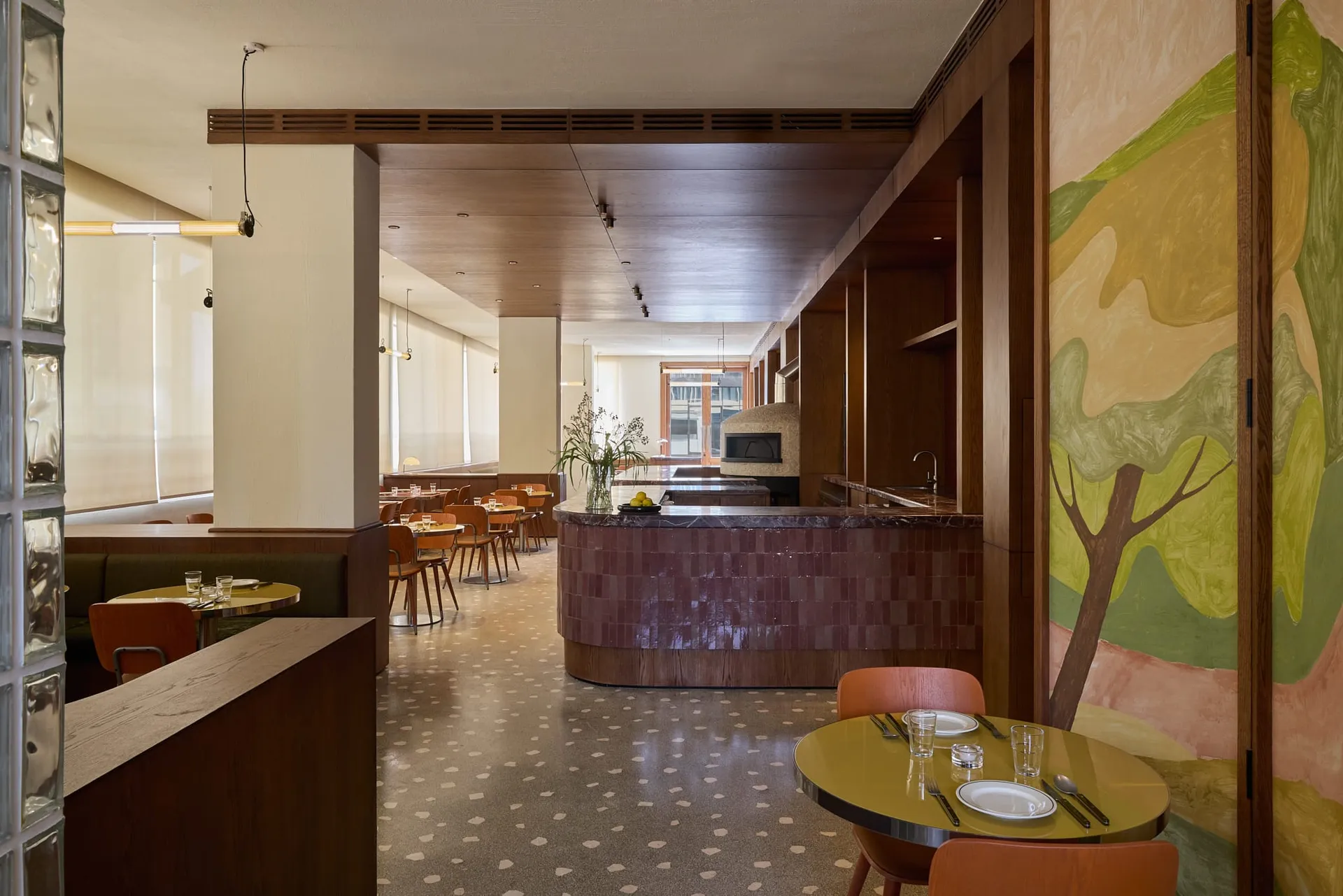 A modern restaurant interior with a dining area featuring a long wooden table and chairs, and a kitchen area with a counter and appliances, lit by soft warm lighting