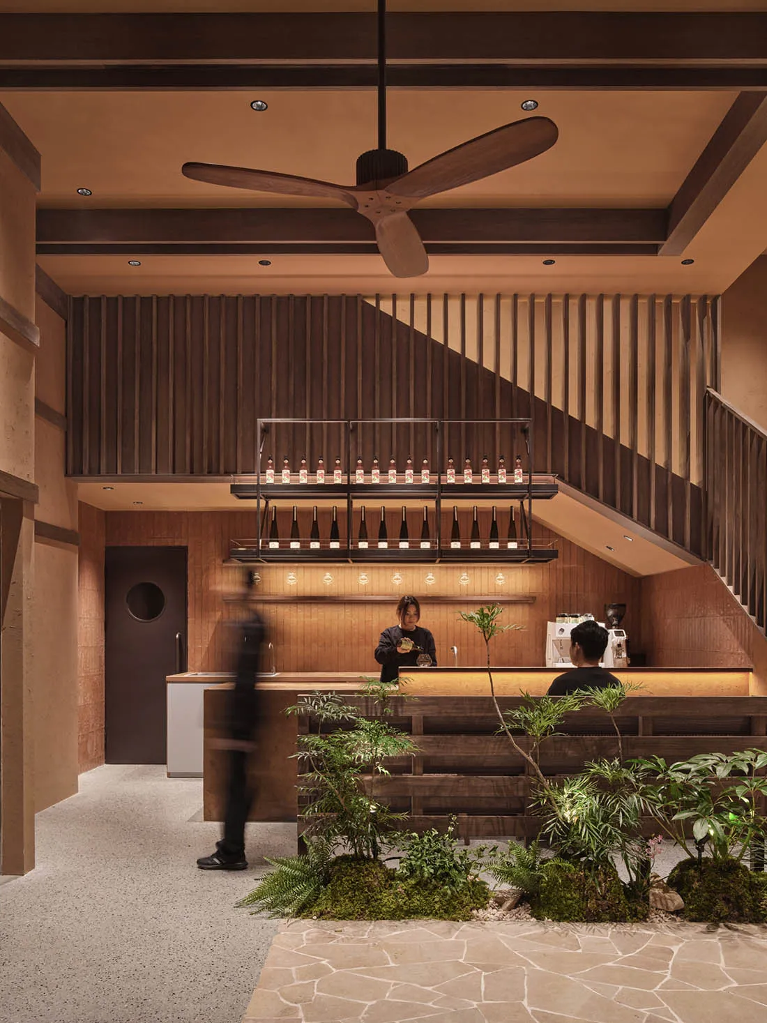 Warm and inviting restaurant interior with a bar, staircase, and potted plants, featuring wood accents and a large fan