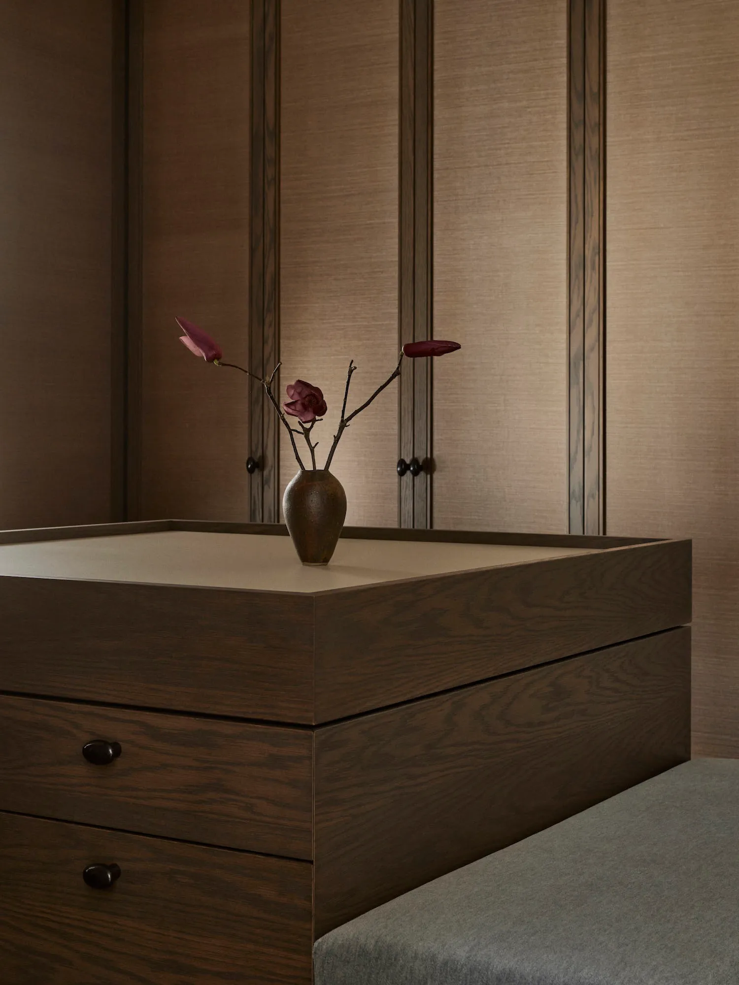 A cozy bedroom with a wooden dresser, a vase of flowers, and a bed in the background, creating a warm and inviting atmosphere