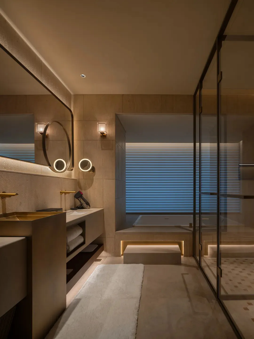 A modern bathroom with a beige color scheme, featuring a large mirror, sink, and bathtub, with a dimly lit atmosphere and a combination of natural and artificial light sources