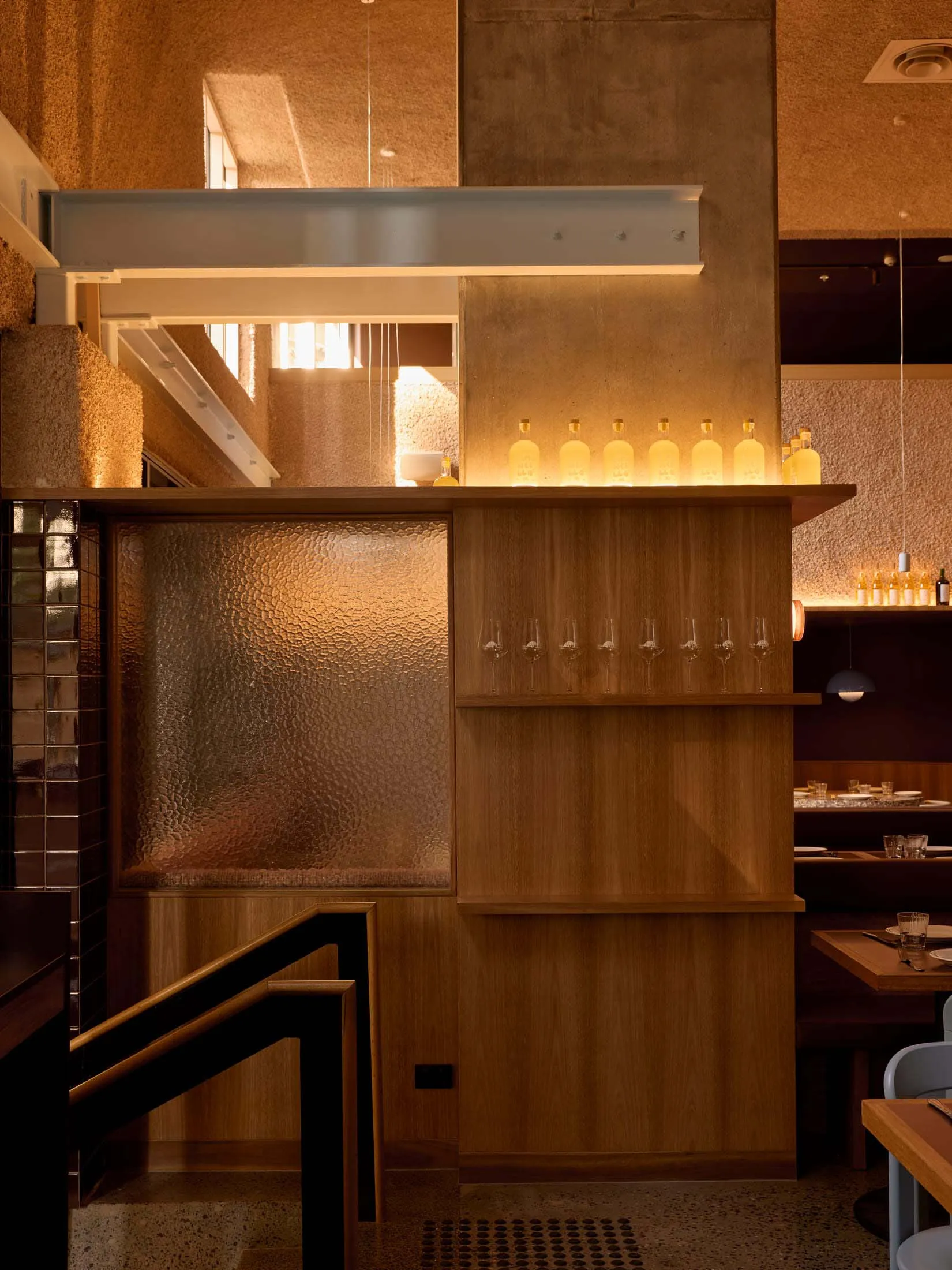 Warm and inviting restaurant with wooden walls, floors, and furniture, featuring a bar with wine glasses and bottles, and natural lighting