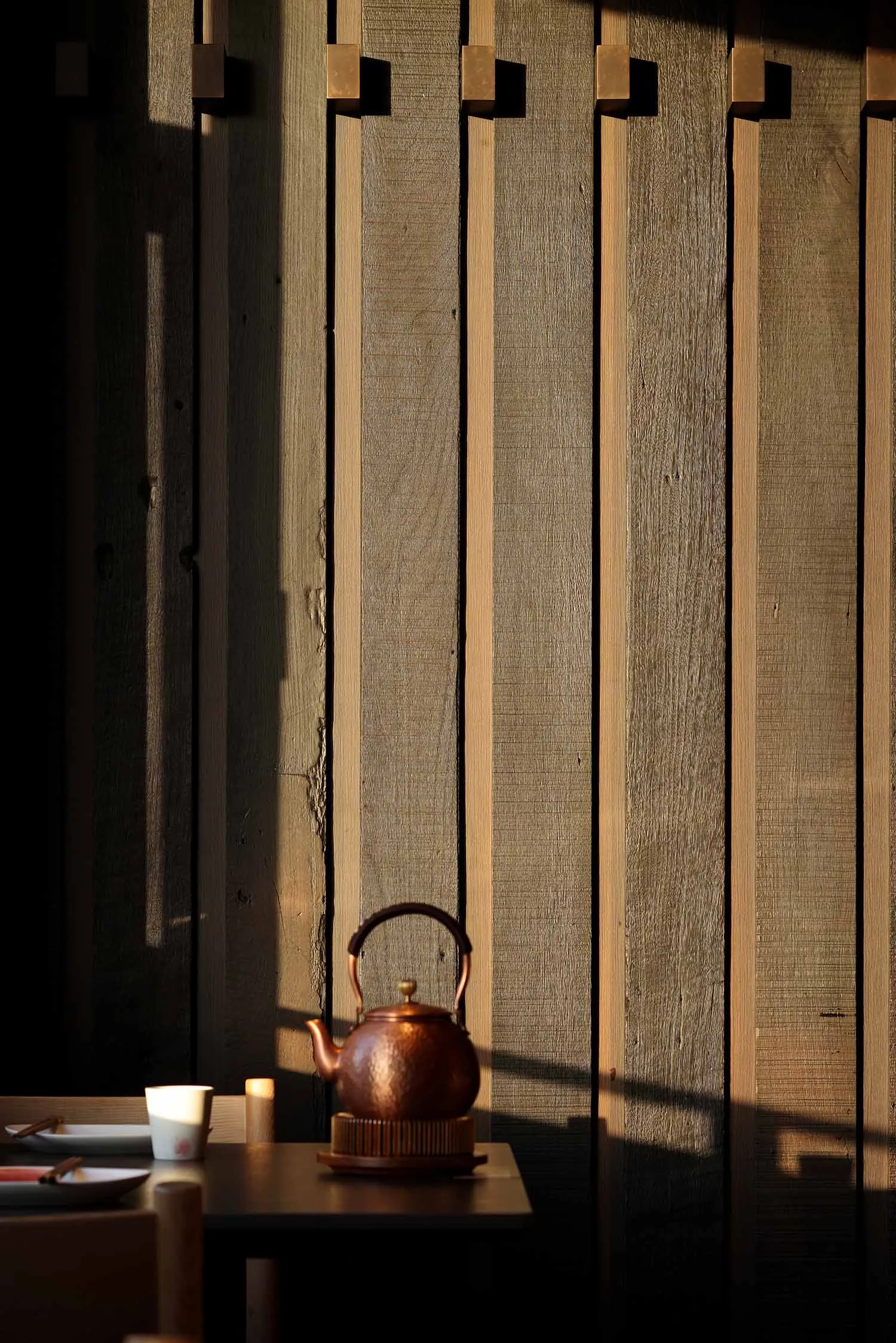A tea kettle and cup sit on a table next to a wooden wall in a warm and inviting indoor setting