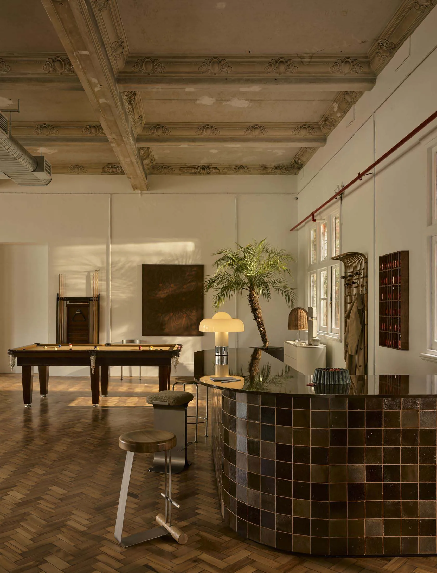 A modern living room with a pool table, bar, and dining table, featuring a tile floor, wood furniture, and a potted plant, with a well-lit and inviting atmosphere