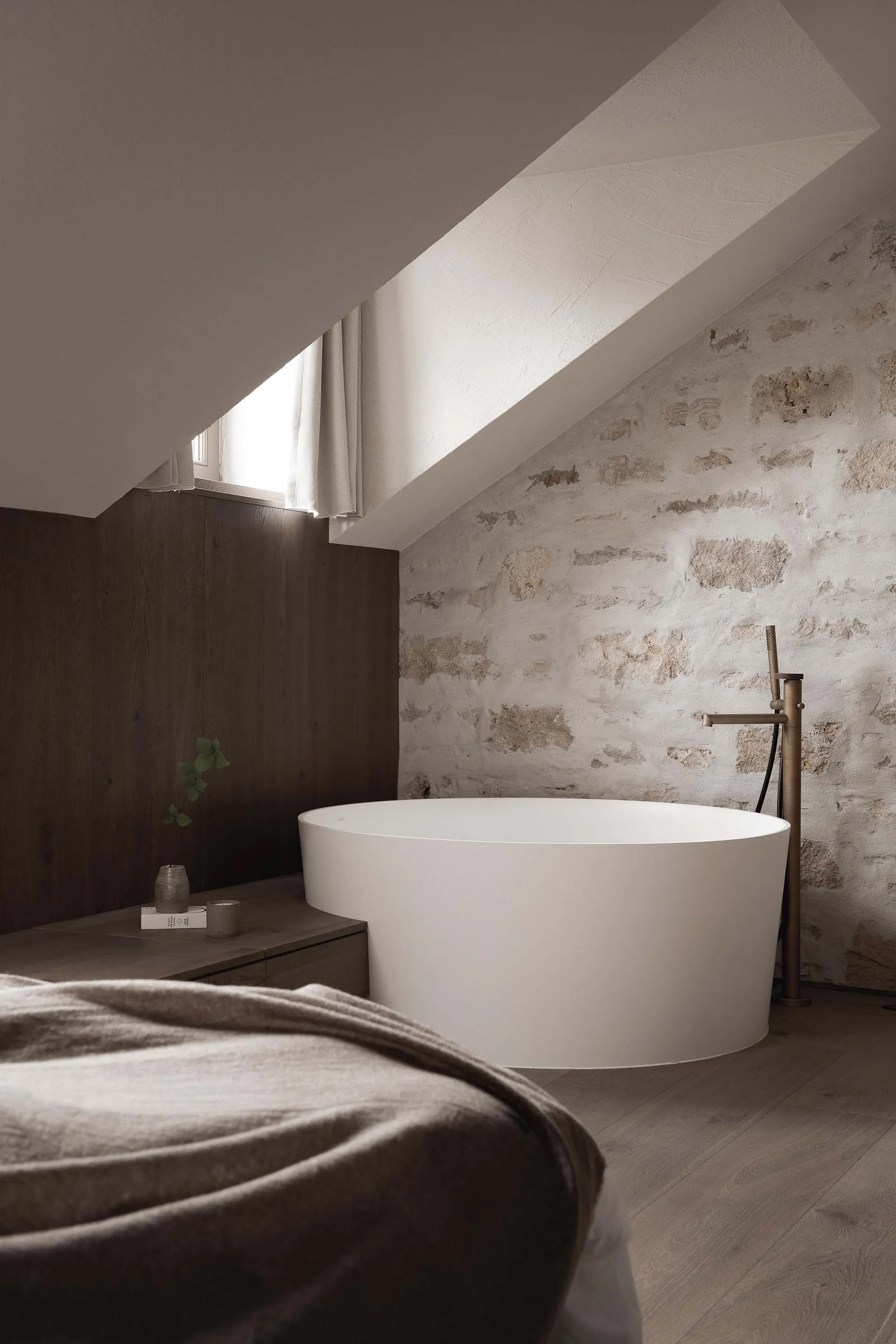 Small bathroom with a white bathtub, sink, and window, set against a stone wall and wooden floor in a dimly lit space