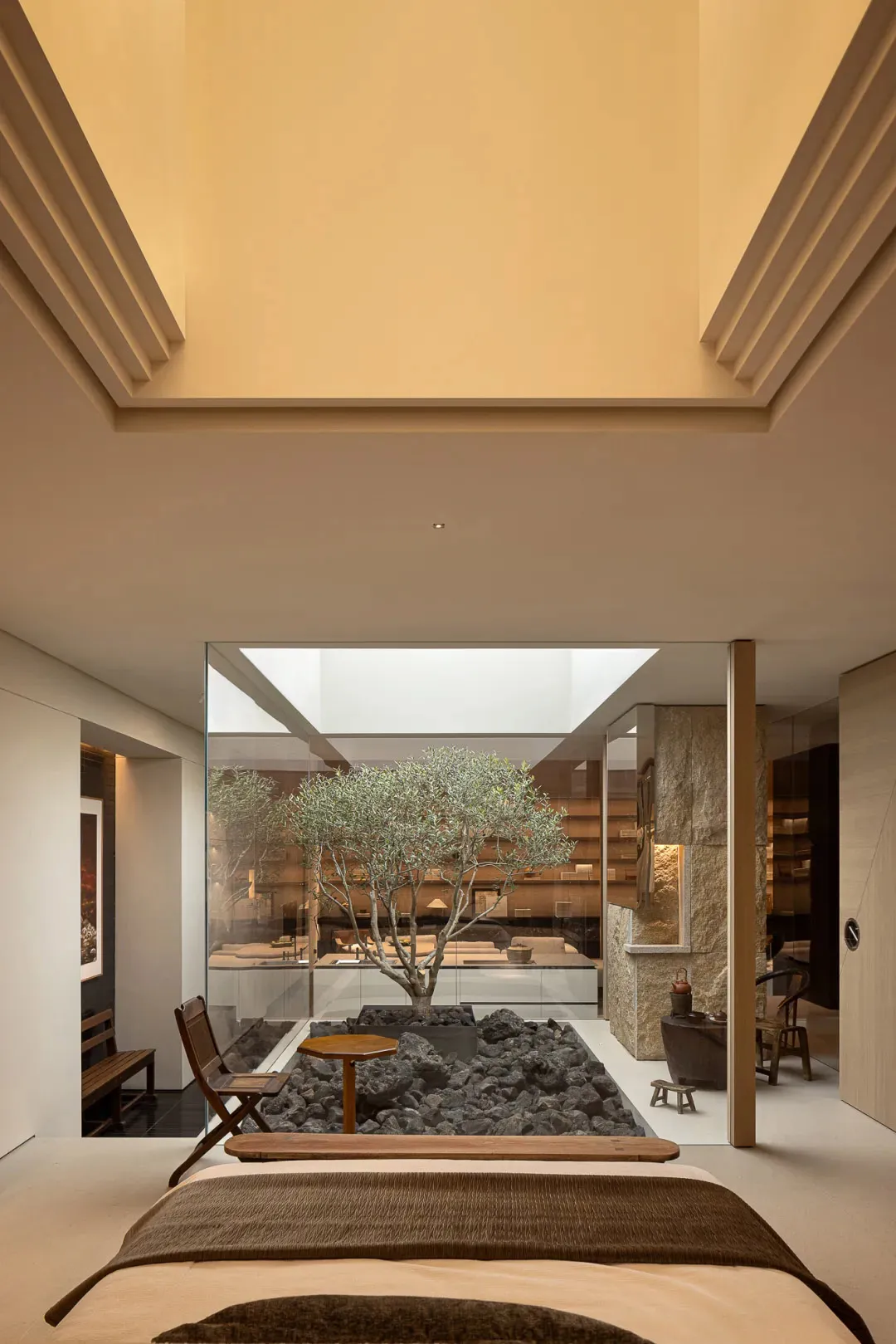 A modern living room with a large tree in the center, surrounded by rocks, a dining table, chairs, a couch, and a potted plant, with a warm and inviting atmosphere created by well-placed lighting