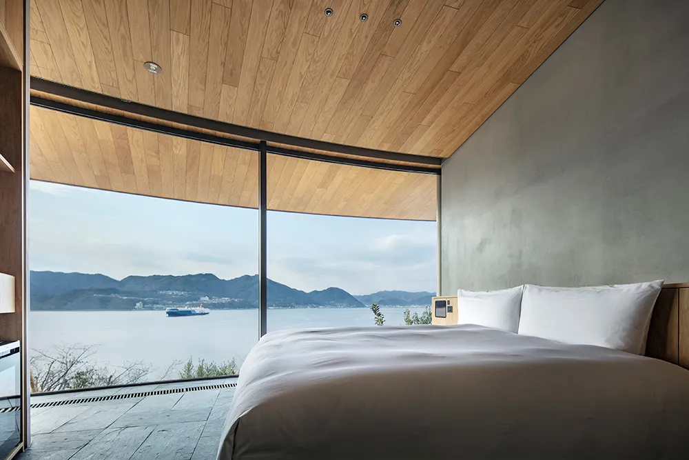 A serene bedroom with a large bed, white bedding, and a view of the ocean through a window, bathed in natural light