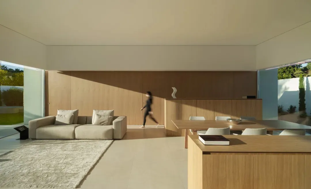 A woman walks towards a dining table in a well-lit living room with a couch and chair