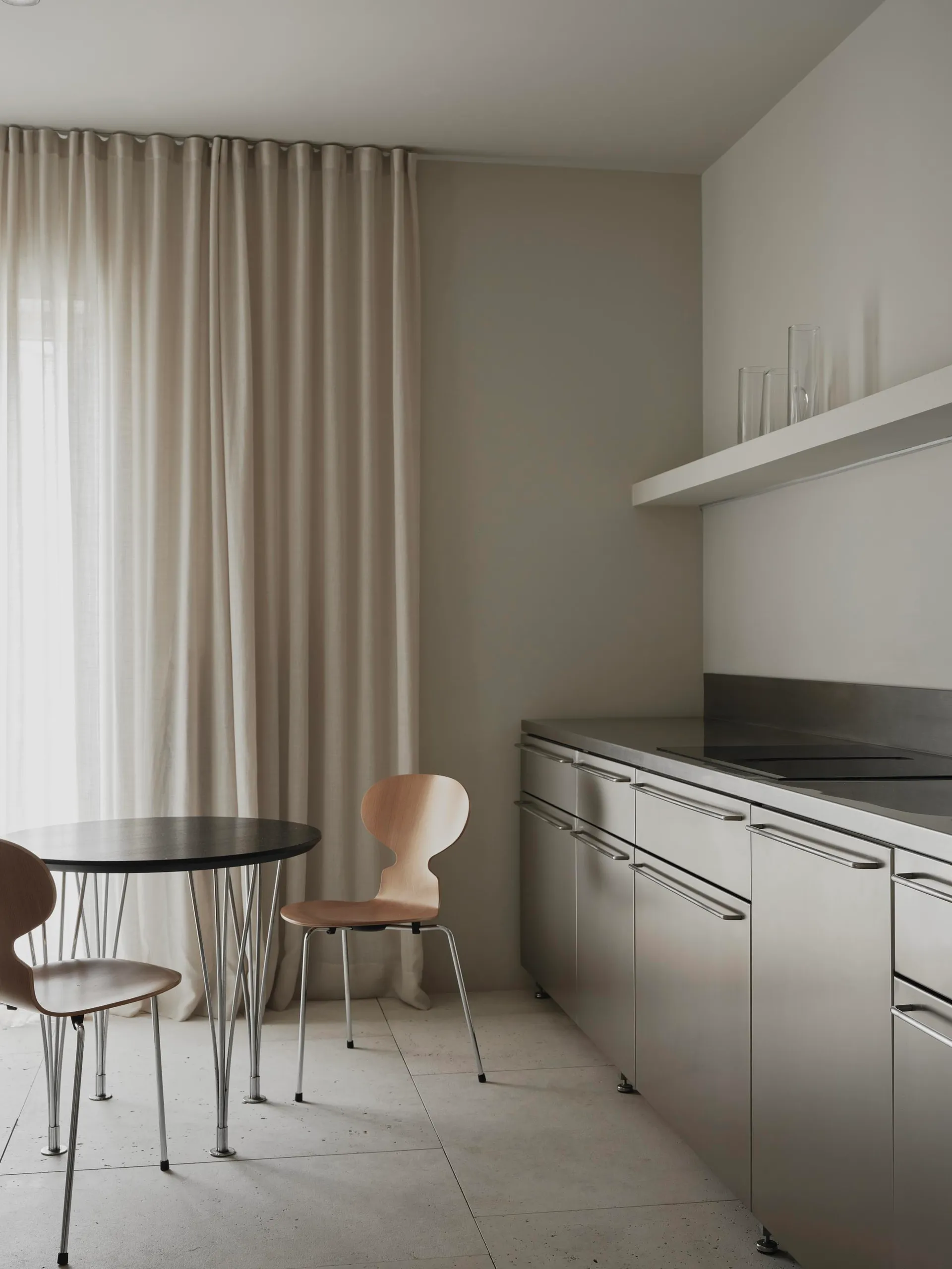 A modern kitchen with a dining table and chairs, surrounded by white surfaces, with natural light and a sink in the background