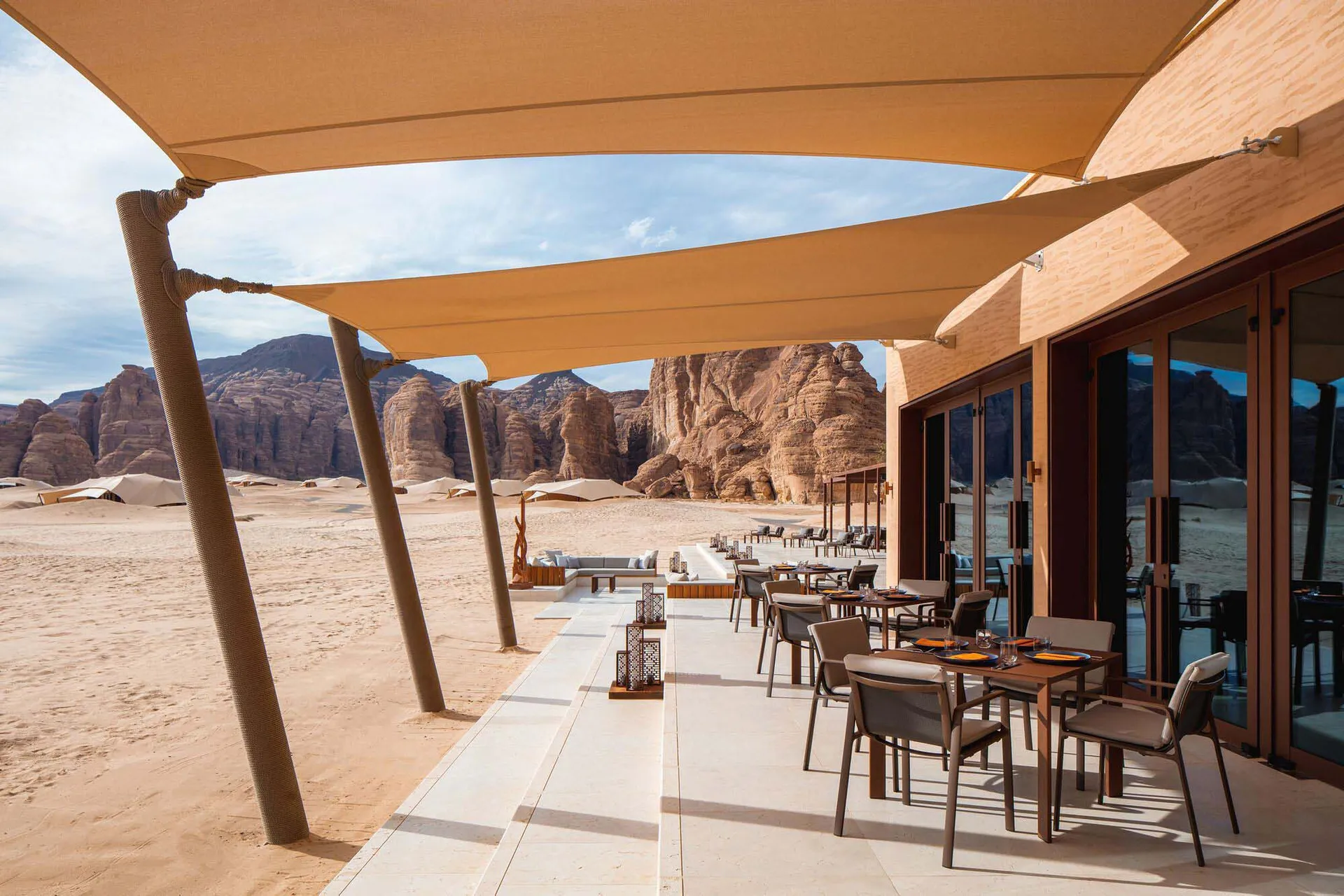 A desert dining area with several chairs and tables arranged under a large umbrella, surrounded by a picturesque desert landscape