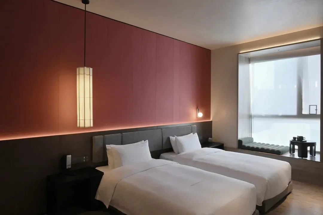 Cozy modern hotel room with two beds, a pink accent wall, and a red wall, featuring white bedding, two lamps, and a comfortable chair near a window with natural light
