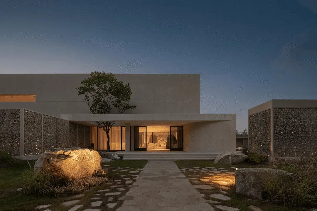 Modern building with a concrete exterior, large window, and door, set against a night sky with a tree in front, illuminated by the building's lights