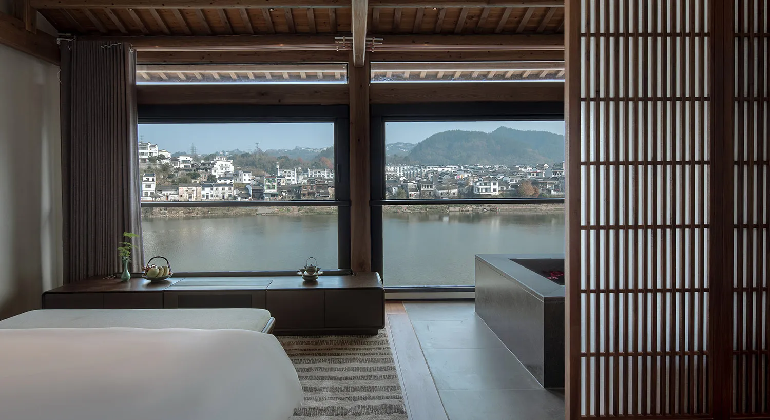 A modern living room with a bed, couch, and sink, featuring a large window with a city view, wood, and glass elements, with natural light and a bright atmosphere