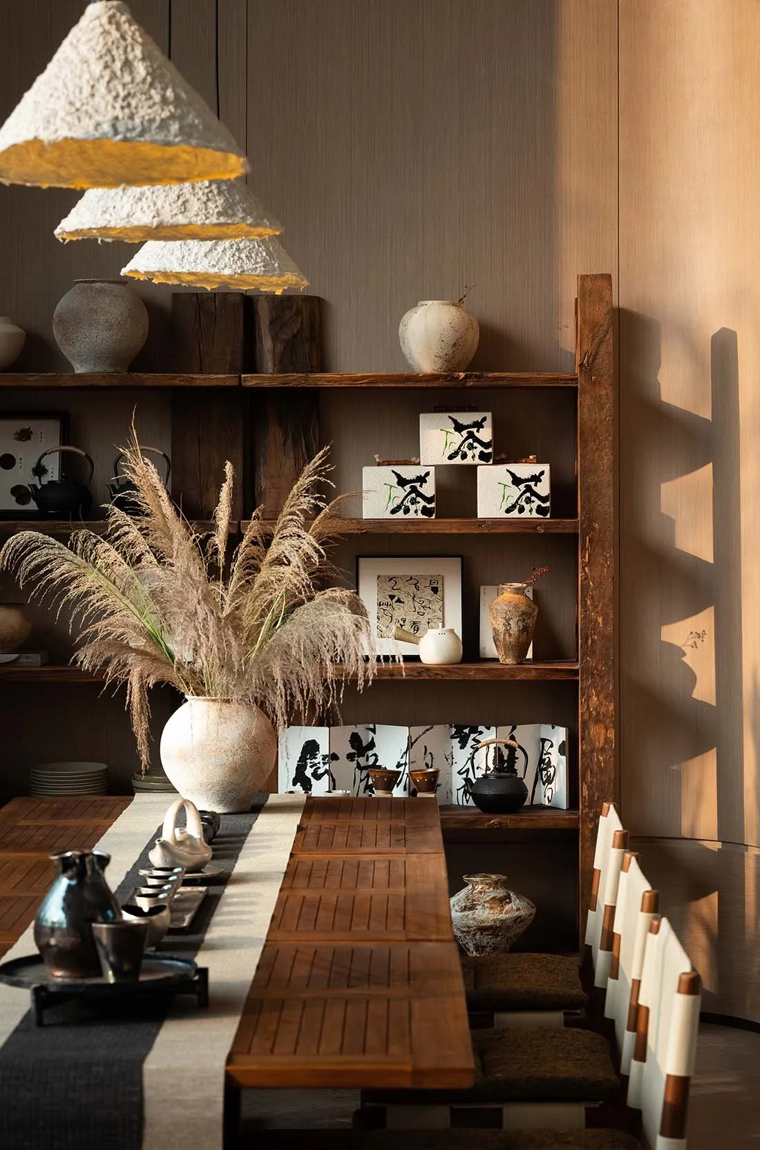 Dining room with a wooden table, chairs, and bookshelf, set with various items and decorated with vases, surrounded by natural light