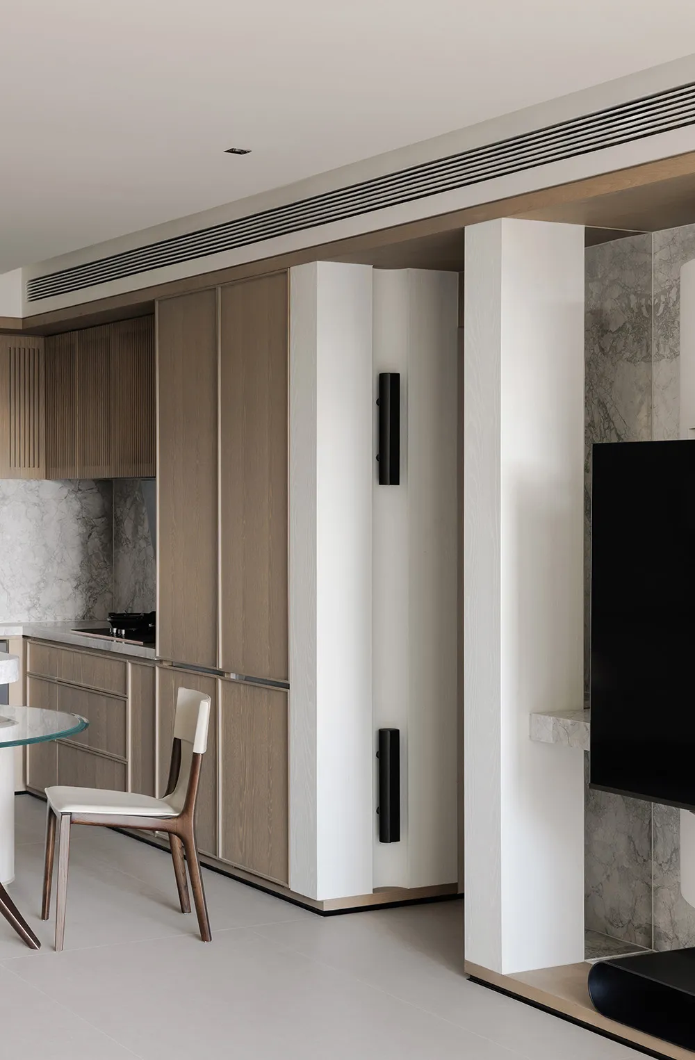 A modern kitchen and living space with wooden cabinets, a glass table, and a black TV, lit by natural light from the windows