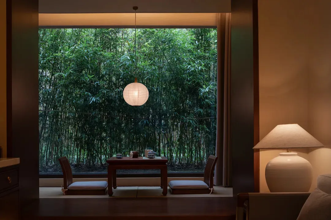 A dining room with a wooden table and chairs, a lamp, and a window with a forest view, creating a warm and inviting atmosphere